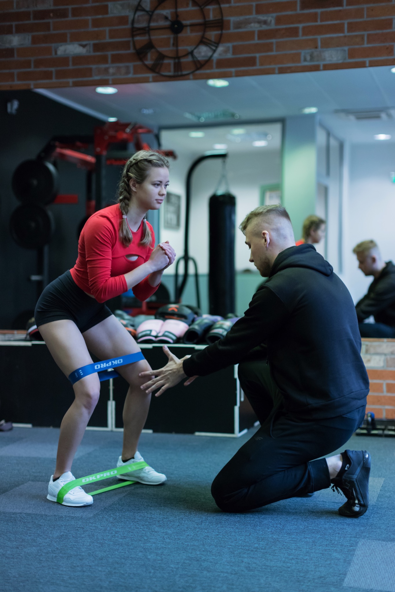 Kobieta w czerwonej koszulce i czarnych spodenkach wykonuje przysiad z gumami oporowymi, nadzorowana przez mężczyznę w czarnej bluzie w siłowni z ceglaną ścianą i workiem treningowym w tle.