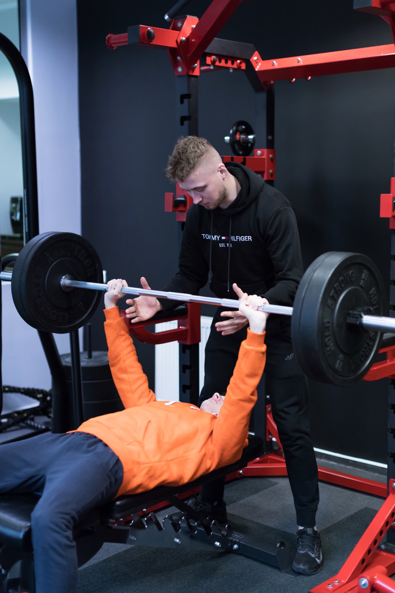 Mężczyzna w pomarańczowej bluzie wyciska sztangę na ławeczce pod okiem instruktora w czarnej bluzie z kapturem w siłowni.