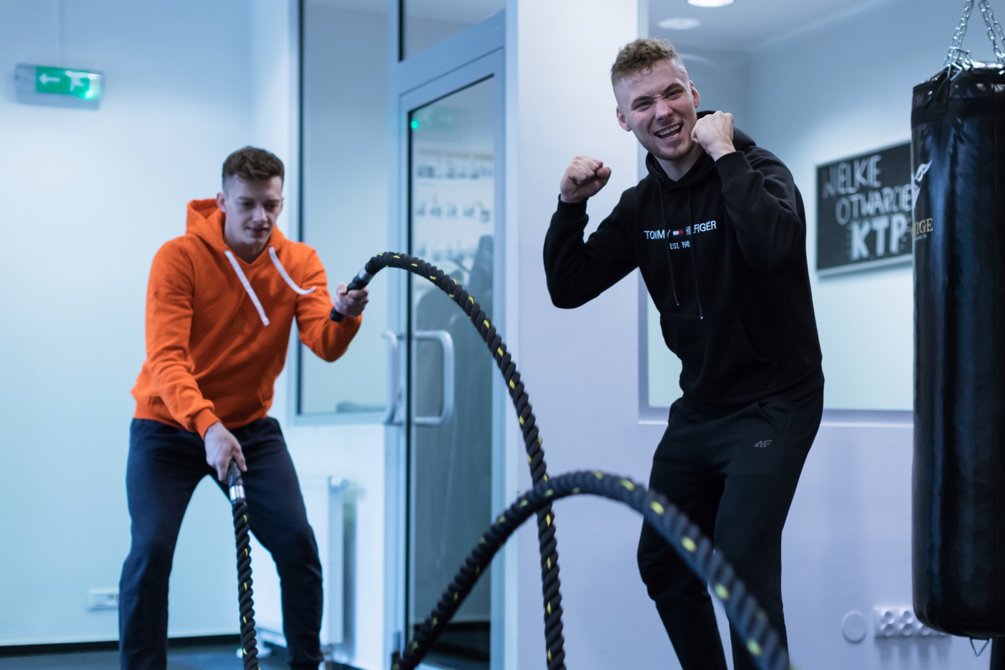 Dwóch mężczyzn ćwiczy na siłowni z linami treningowymi, jeden w pomarańczowej bluzie wykonuje ćwiczenie, drugi w czarnej bluzie z uniesionymi pięściami w geście radości, w tle worek treningowy...