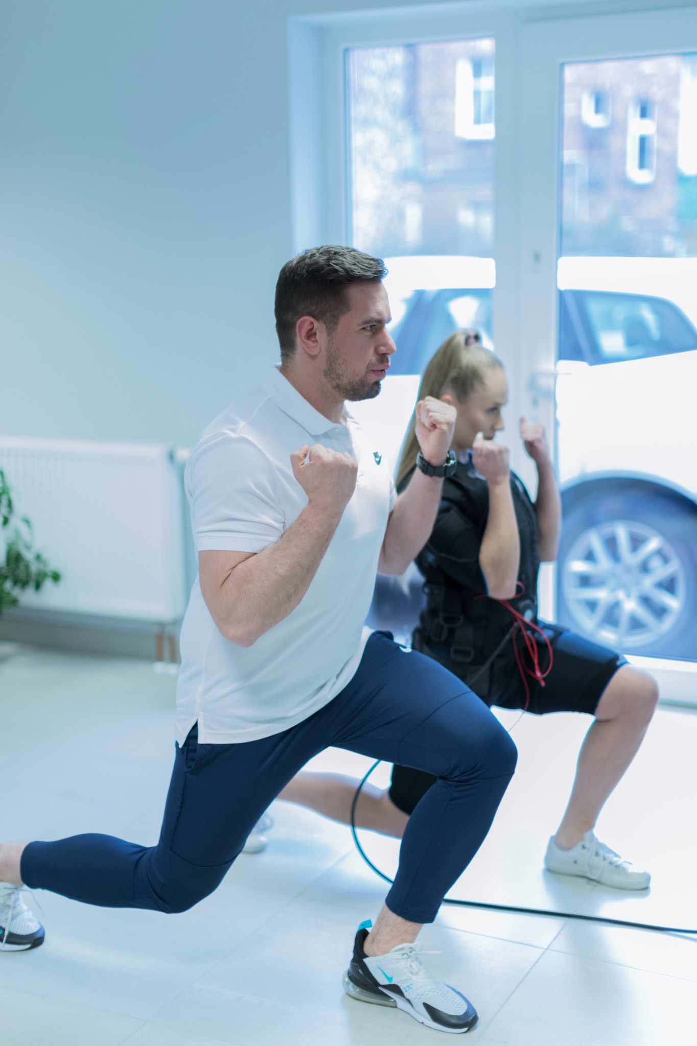 Mężczyzna w białej koszulce polo i granatowych spodniach wykonuje wypad do przodu podczas treningu EMS; kobieta w elektrostymulacji również ćwiczy w tle, widoczna w tle biała ściana i okno...