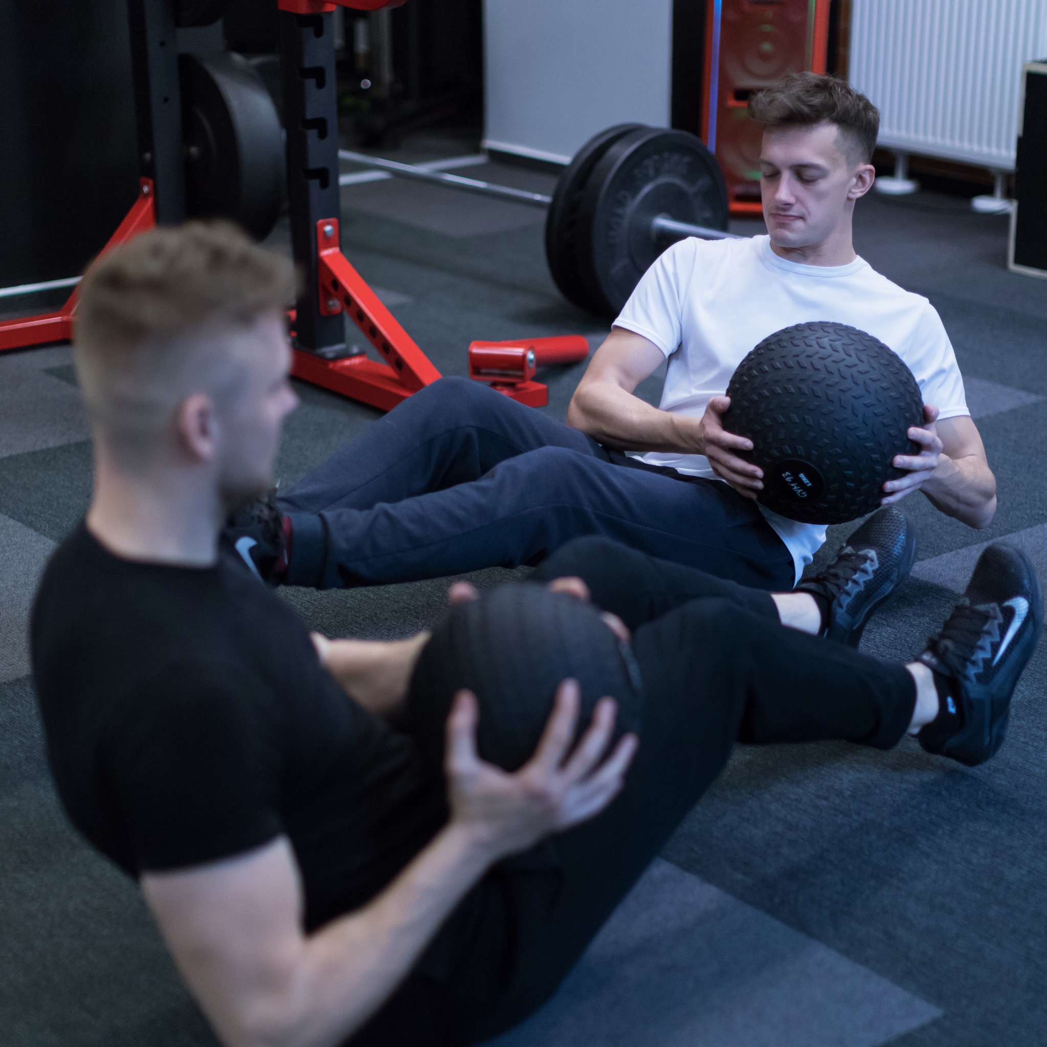 Dwóch mężczyzn wykonuje ćwiczenia z piłkami lekarskimi w siłowni, z widocznym sprzętem do podnoszenia ciężarów w tle.