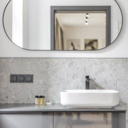 PIK ARCHITEKTURA MARIUSZ ZAŃKO - Nowoczesna łazienka z owalnym lustrem, białą umywalką nablatową i szarą armaturą na tle ściany z szarego kamienia.