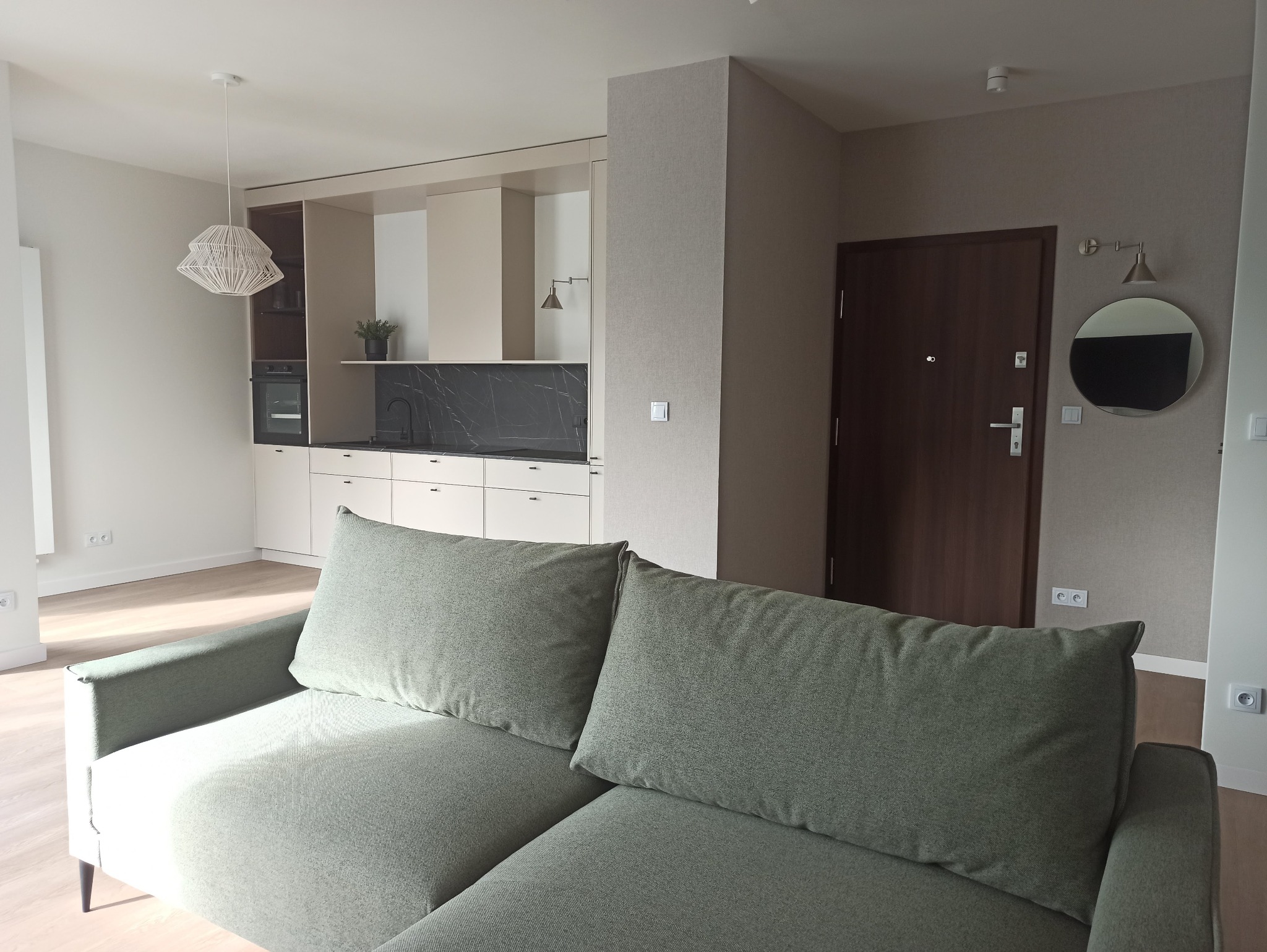 Jasne wnętrze mieszkania z widoczną kuchnią z czarnym blatem, minimalistycznym oświetleniem i zieloną sofą na pierwszym planie.