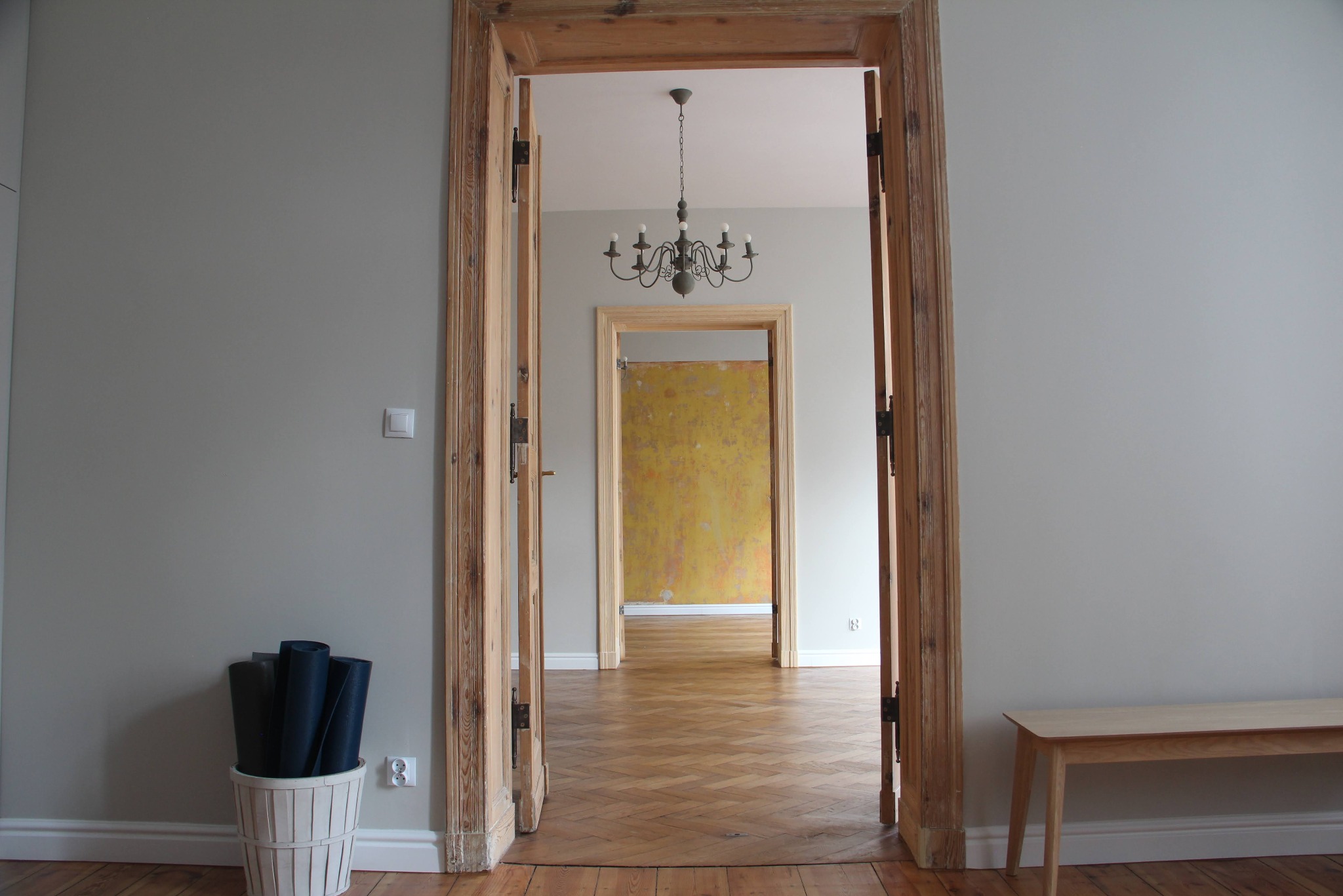 Wnętrze mieszkania po remoncie, widok na otwarte, drewniane drzwi, parkiet w jodełkę, szara ściana, minimalistyczna ławka i kosz z czarnymi matami, w tle widoczny żyrandol i fragment pomalowanej...