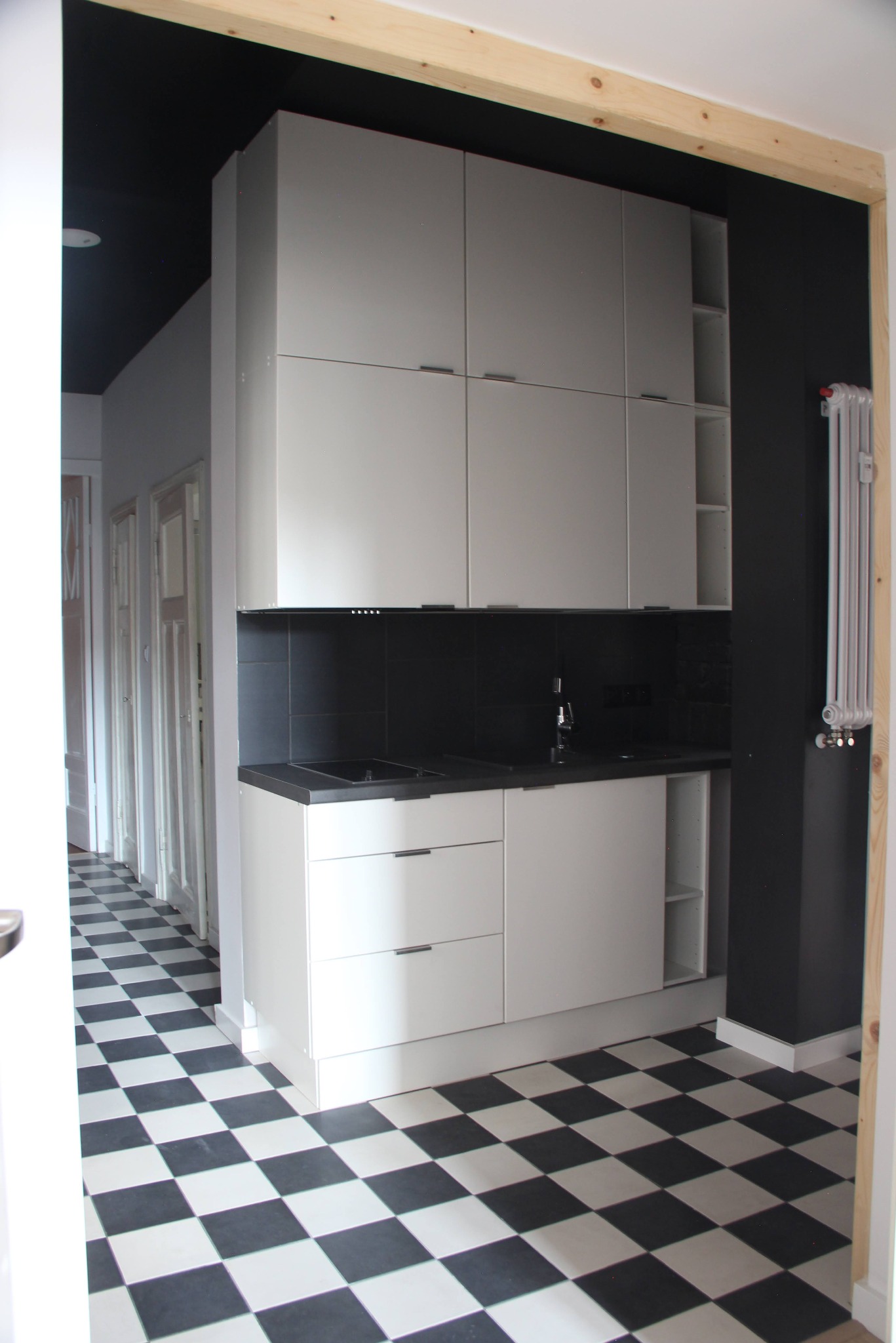 Nowoczesna kuchnia z białą zabudową szafek i czarnym blatem, czarno-biała podłoga w szachownicę, czarne ściany i sufit w tle, widok z korytarza przez framugę drzwi.