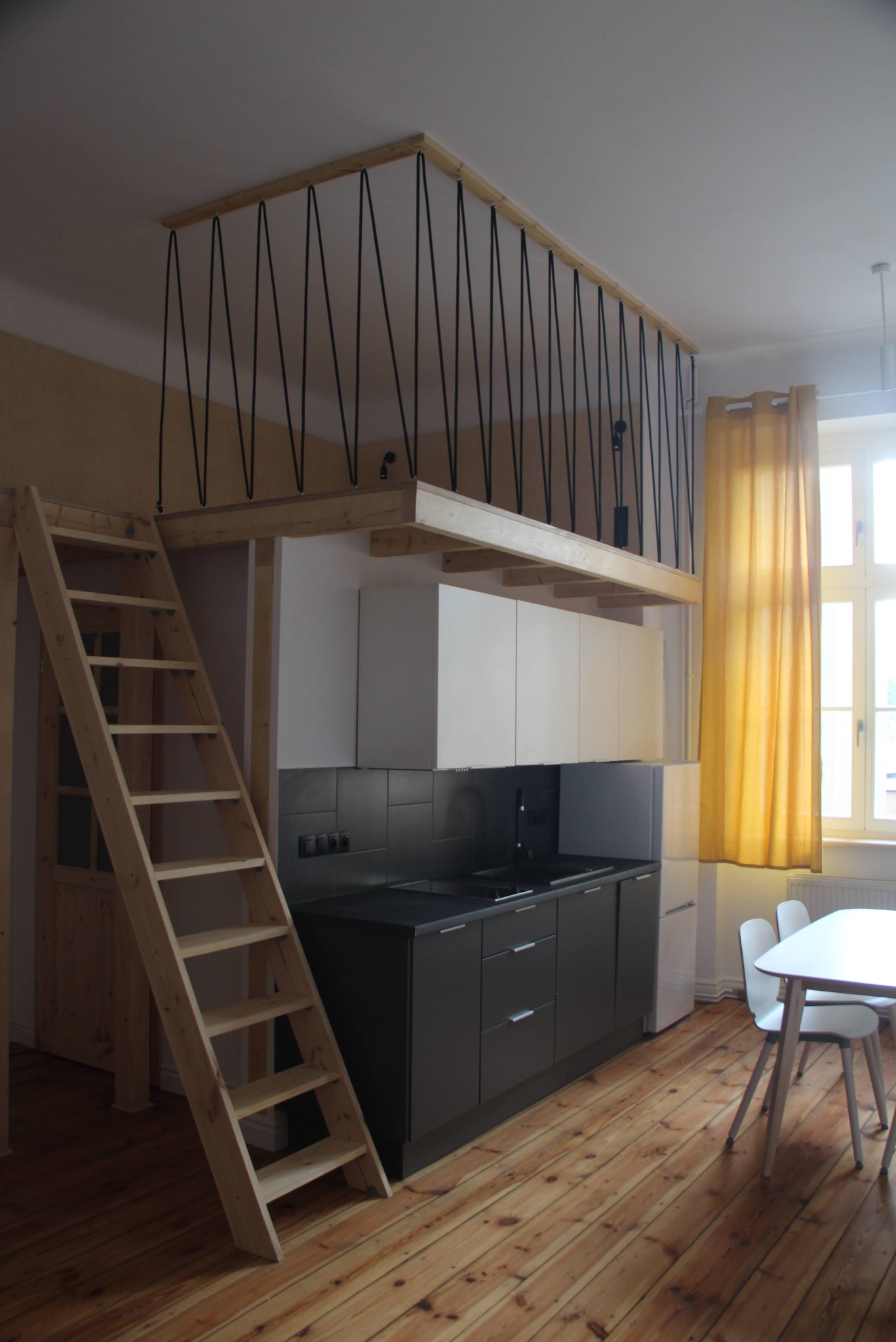 Nowoczesna kuchnia z antresolą zabezpieczoną czarnymi linkami, drewniane schody, parkiet i stół z krzesłami przy oknie z żółtą zasłoną.