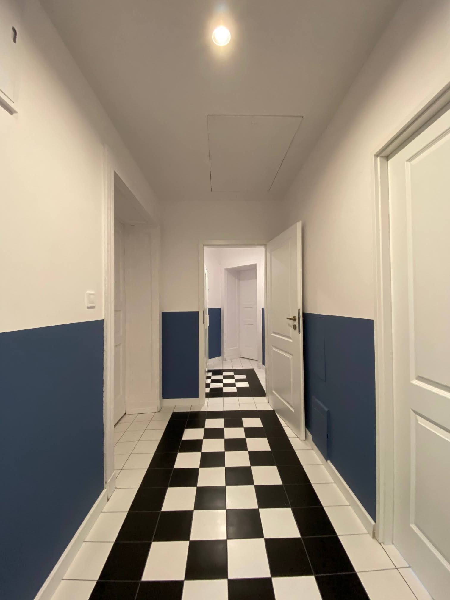 Korytarz z biało-czarną szachownicą na podłodze, ściany pomalowane w połowie na biało, w połowie na granatowo, białe drzwi otwarte do kolejnych pomieszczeń.