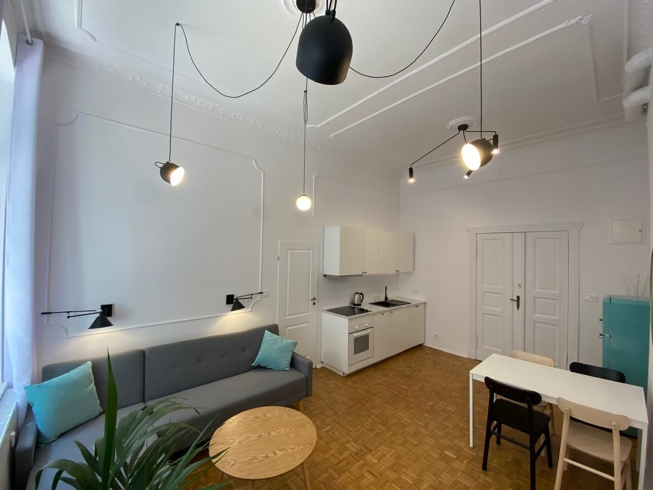 Jasne wnętrze mieszkania z szarą sofą, drewnianym stolikiem kawowym, białymi meblami kuchennymi i nietypowym oświetleniem sufitowym.