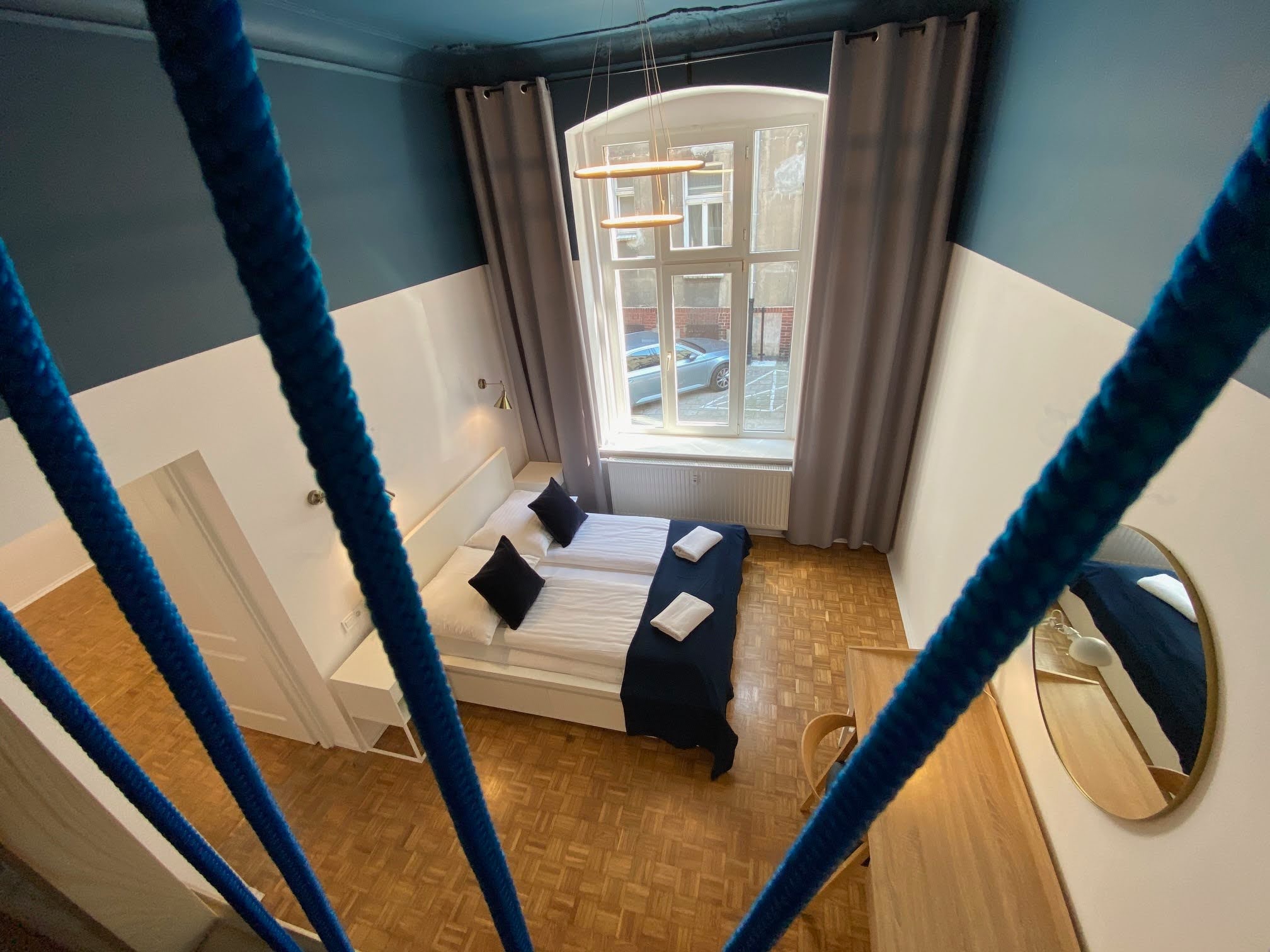 Widok z góry na pokój z dwoma łóżkami z niebieskimi akcentami kolorystycznymi, widoczny przez niebieskie liny balustrady schodów, parkiet na podłodze, białe ściany do połowy wysokości, niebieska...