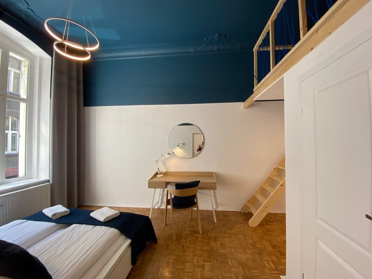 Pokój z antresolą, biurkiem i łóżkiem, ściany pomalowane na biało i granatowo, parkiet w jodełkę, nowoczesna lampa wisząca.