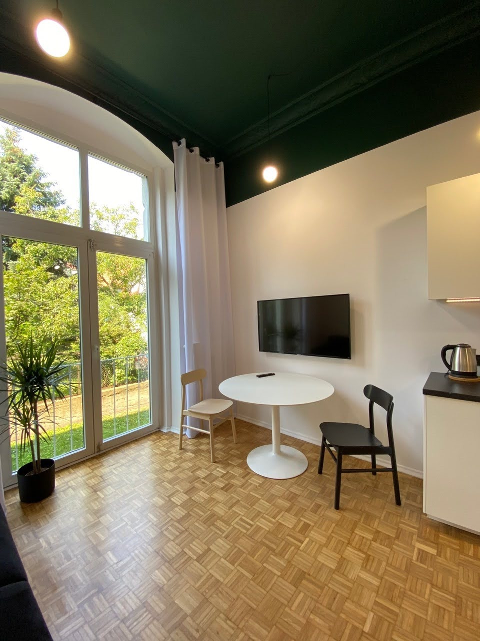 Wnętrze mieszkania po remoncie: drewniana podłoga parkietowa, białe ściany, ciemnozielony sufit, okno balkonowe z widokiem na zieleń, okrągły biały stół, dwie krzesła (jasne i czarne), telewizor...