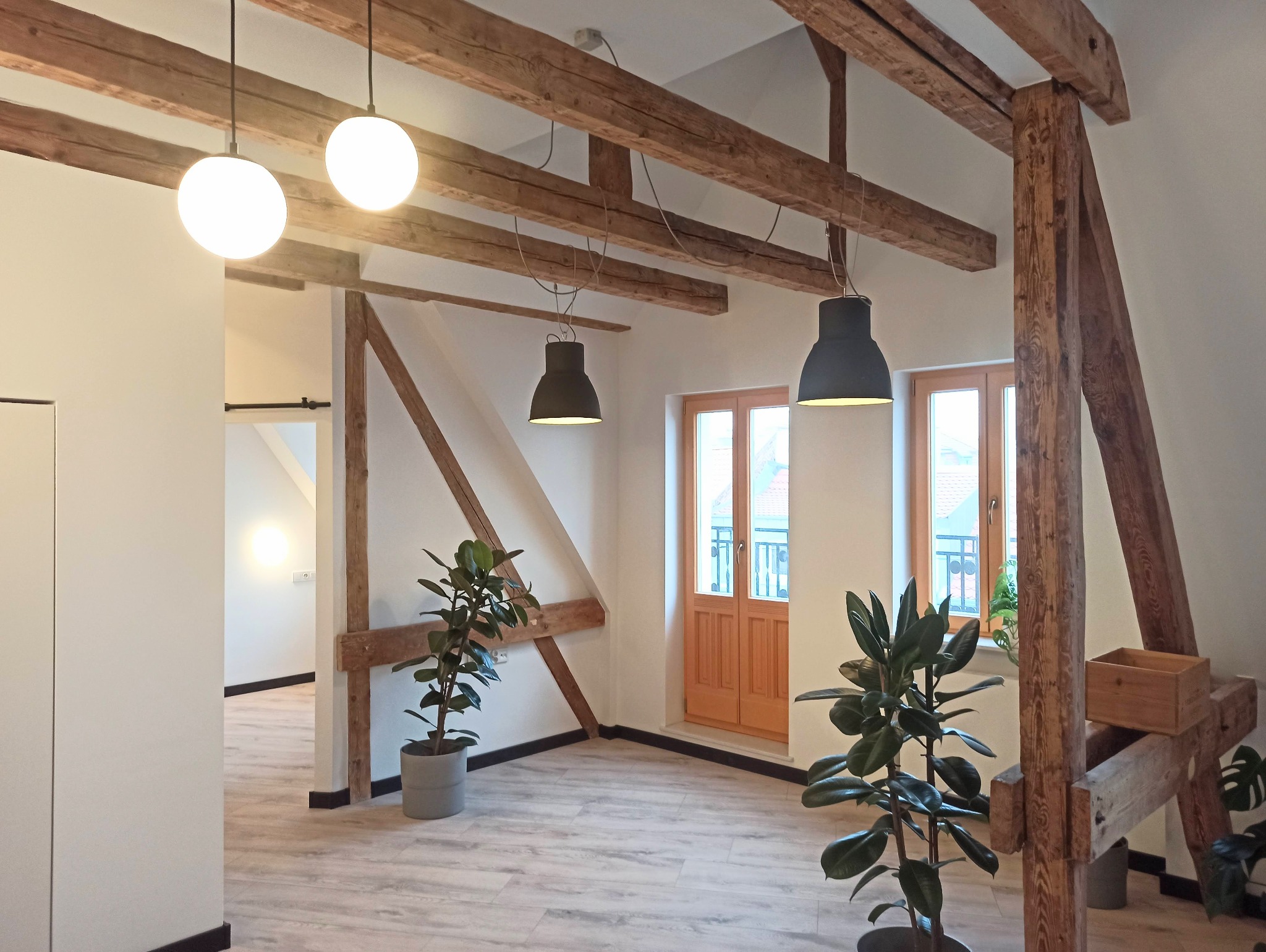 Jasne, przestronne poddasze z drewnianymi belkami stropowymi, białymi ścianami, podłogą z paneli imitujących drewno, dwuskrzydłowymi drewnianymi drzwiami, nowoczesnymi lampami wiszącymi i roślinami...