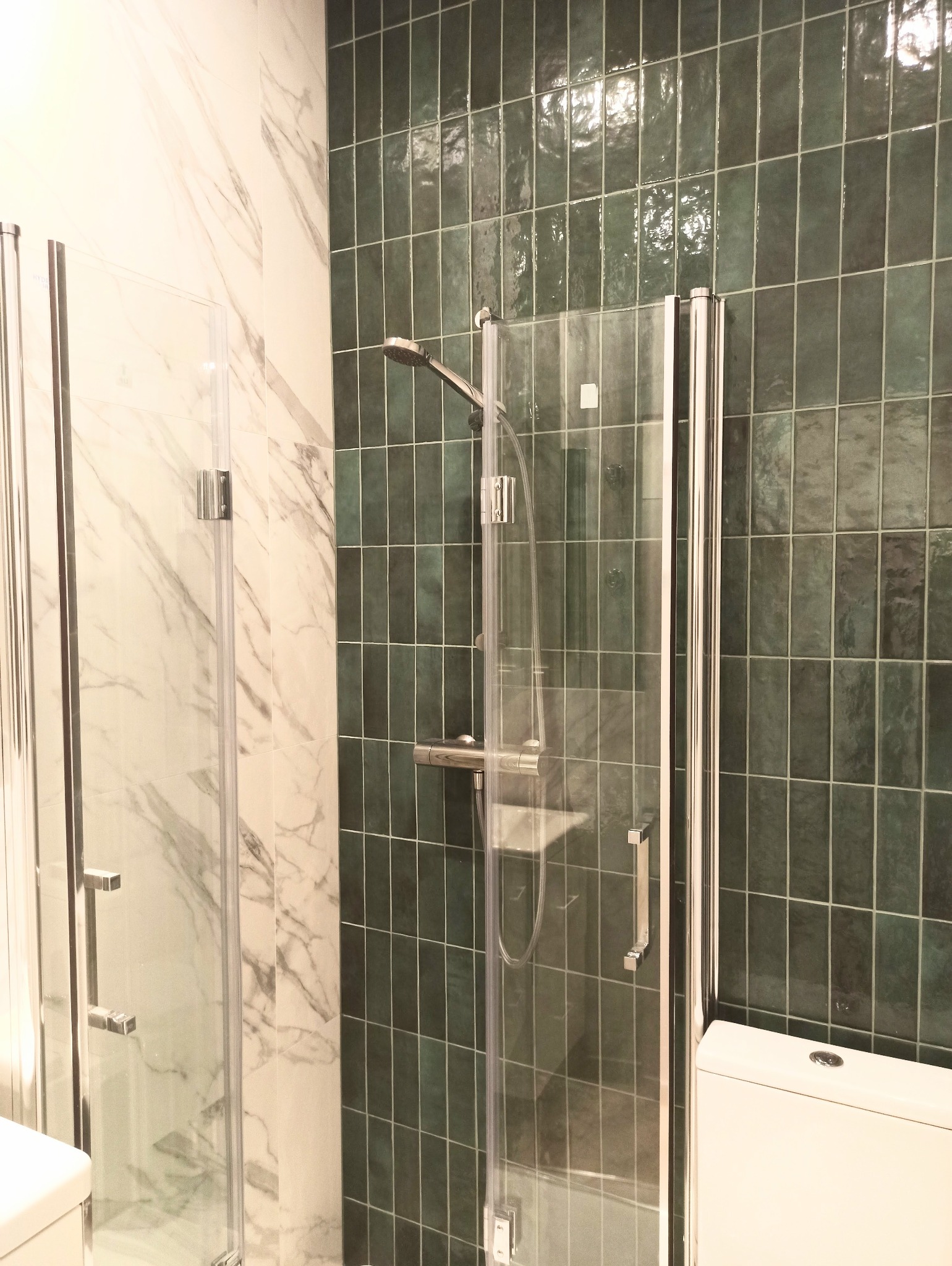 Nowoczesna kabina prysznicowa z transparentnymi drzwiami, zielone, błyszczące płytki ścienne ułożone pionowo, oraz marmurowe płytki na ścianie obok, widoczny fragment toalety.