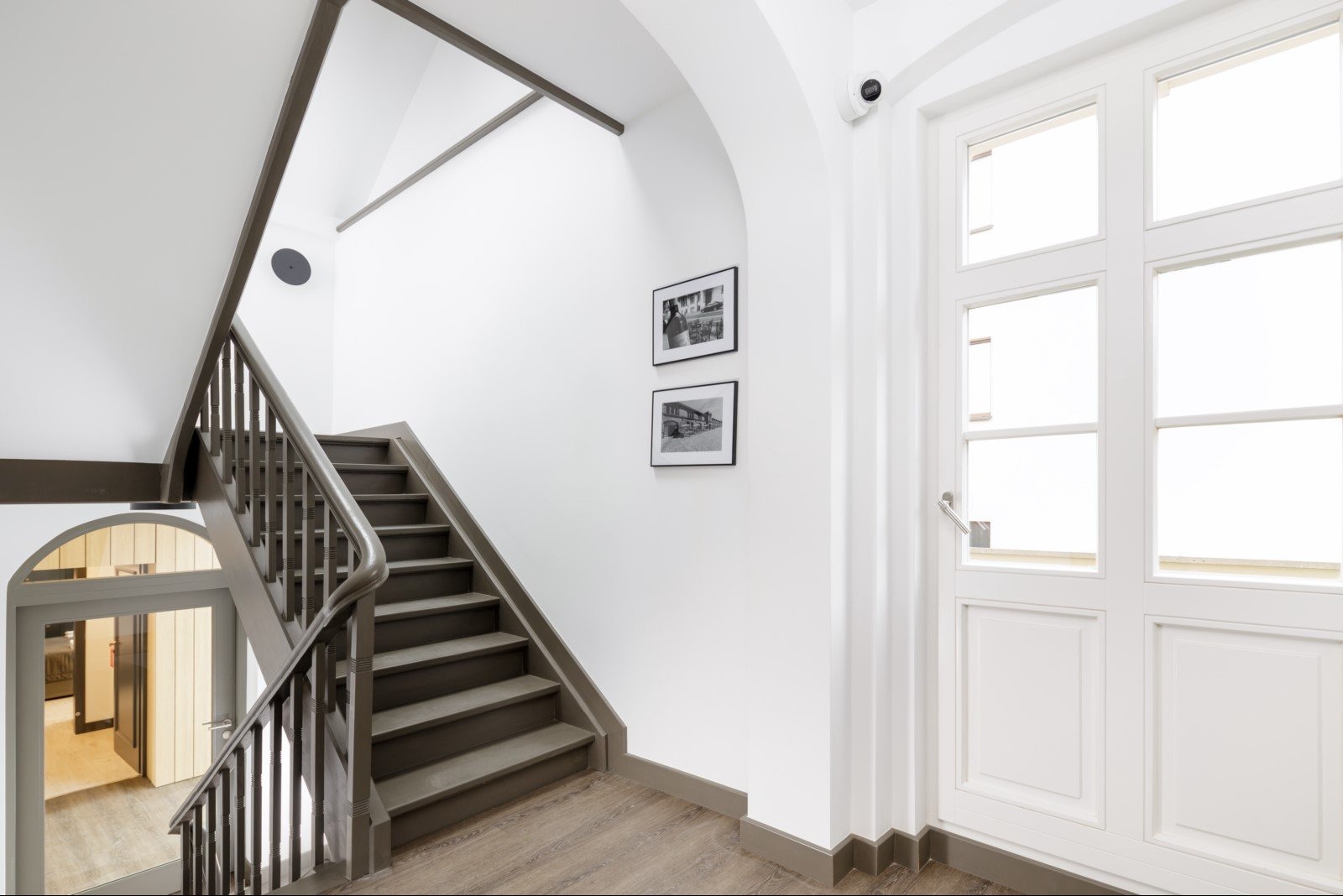 Klatka schodowa w jasnych barwach z drewnianymi schodami i białymi ścianami, ozdobiona dwoma czarno-białymi zdjęciami w ramkach. Widoczna biała, przeszklona wewnętrzna brama oraz kamera...