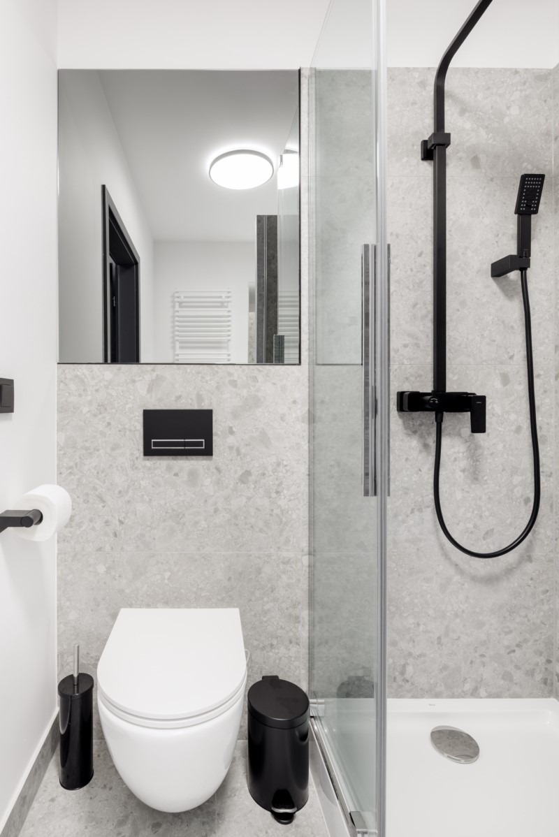 Nowoczesna łazienka wykończona szarym kamieniem: wisząca toaleta, czarne akcesoria (szczotka, kosz, uchwyt na papier), minimalistyczna kabina prysznicowa z czarną armaturą, duże lustro bez ramy.