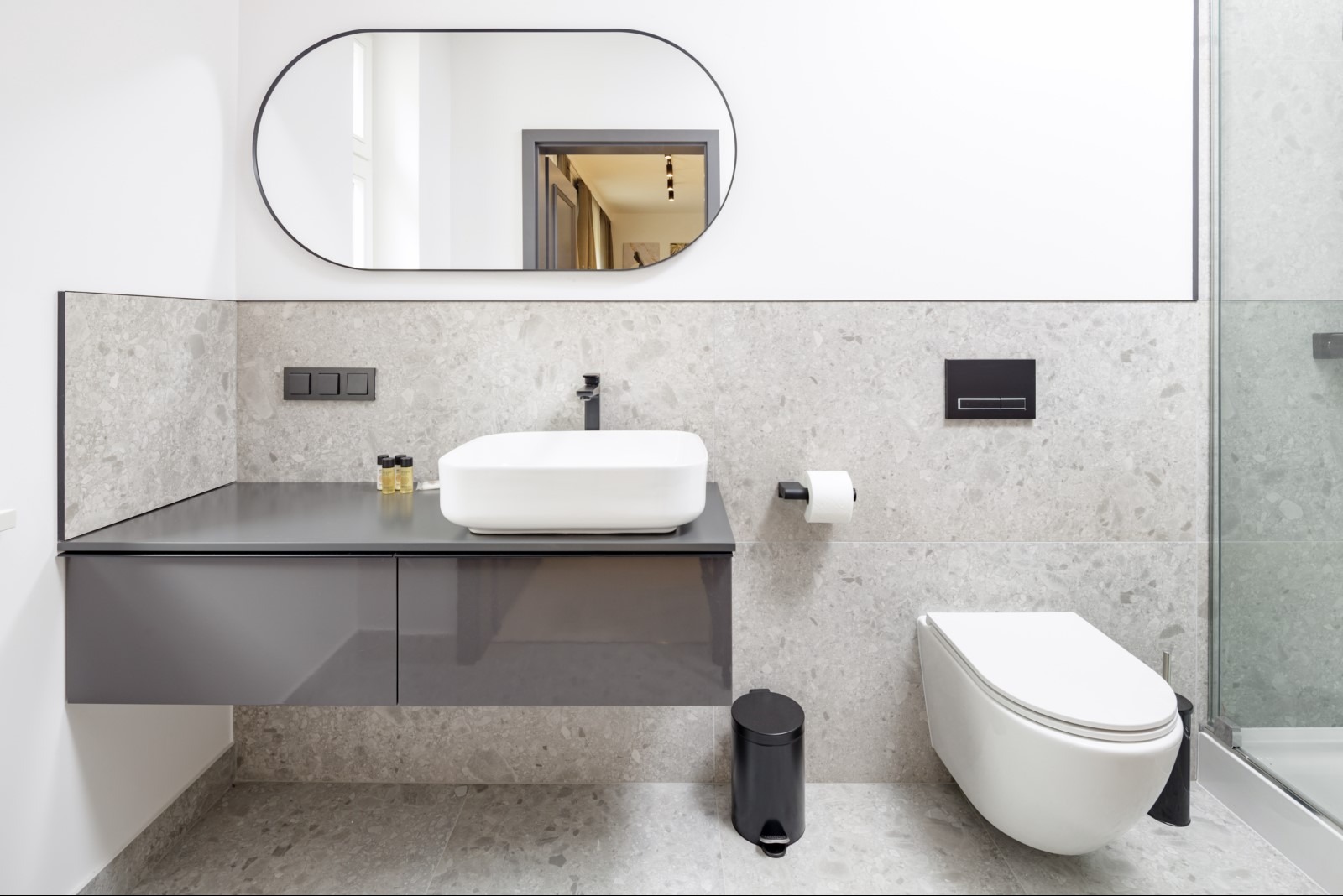 Nowoczesna łazienka z owalnym lustrem, podwieszaną szafką z umywalką, wiszącą toaletą i szarymi płytkami imitującymi kamień.