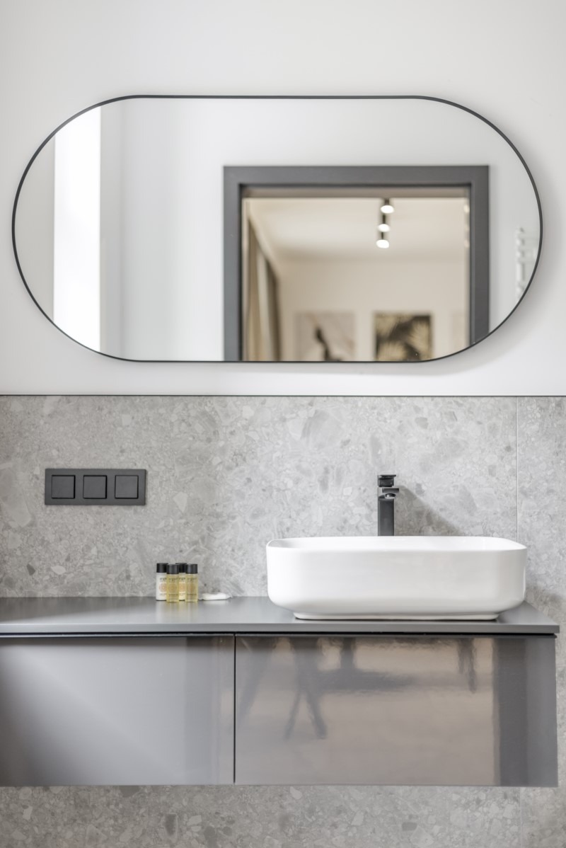 Nowoczesna łazienka z owalnym lustrem, białą umywalką nablatową i szarą armaturą na tle ściany z szarego kamienia.