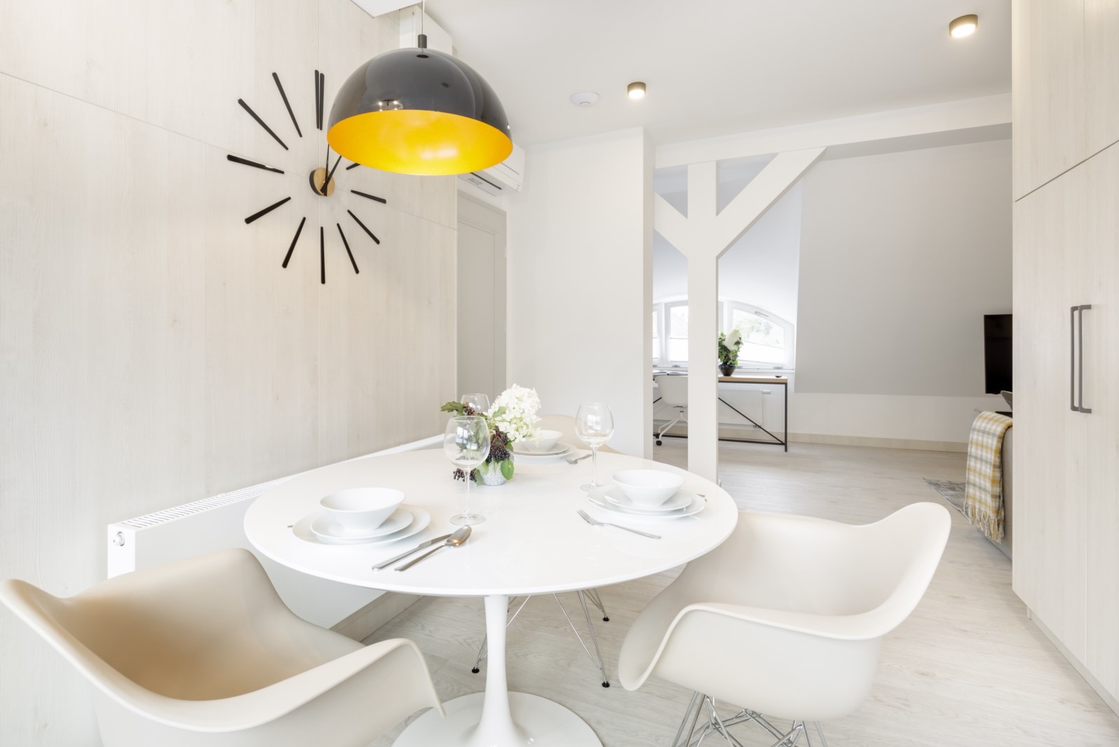 Jasne wnętrze jadalni z okrągłym białym stołem, nakrytym dla dwóch osób, z modernistycznym zegarem na ścianie i widokiem na pokój dzienny w tle.