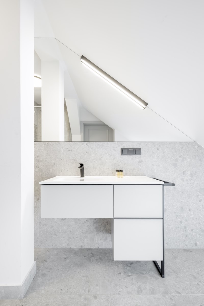 Nowoczesna łazienka w odcieniach szarości i bieli: wisząca szafka z umywalką, czarna bateria, podświetlane lustro na skośnej ścianie poddasza.