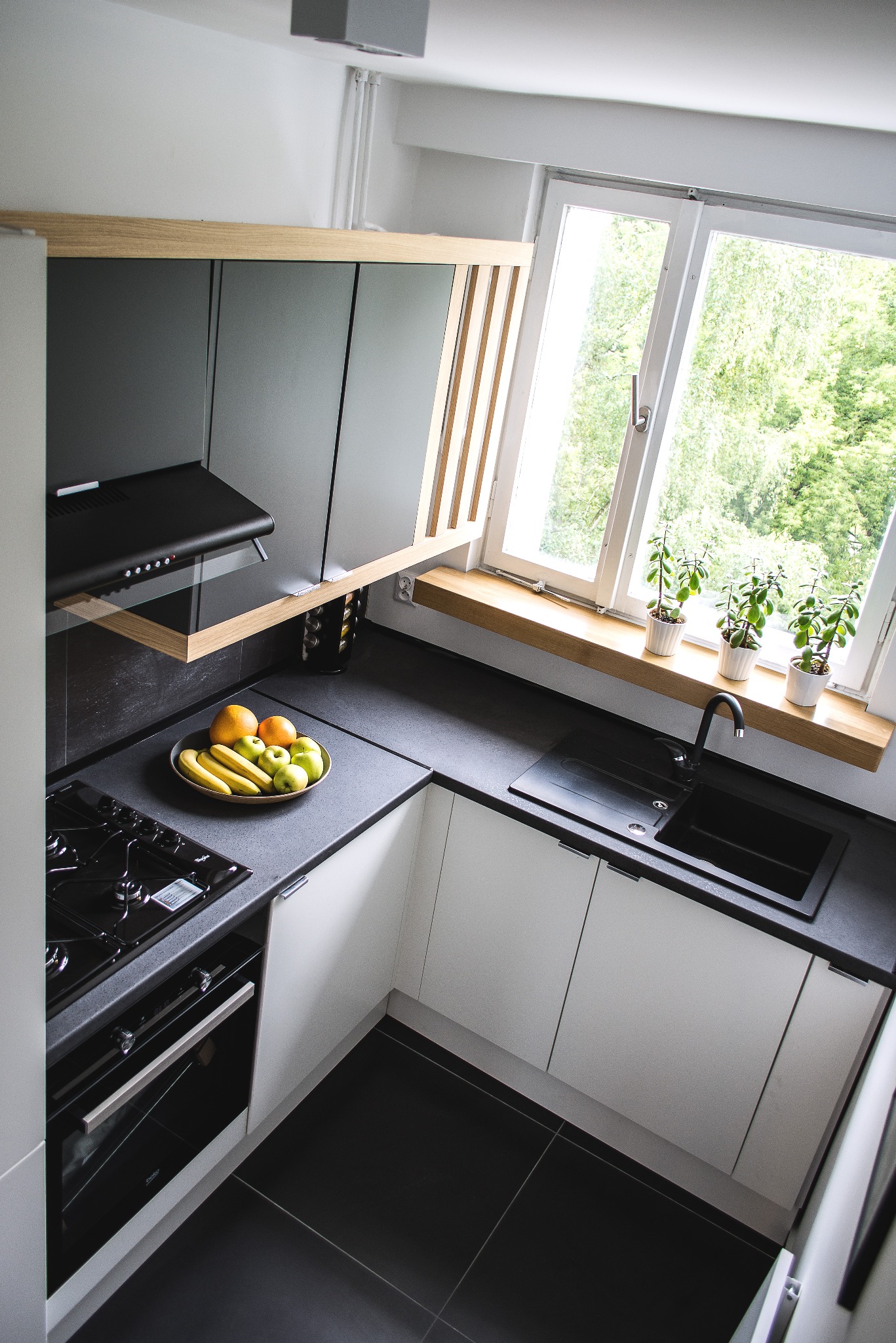 Nowoczesna kuchnia w tonacji bieli i grafitu z czarnymi akcentami, widok z góry, na blacie misa z owocami, za oknem zieleń drzew.