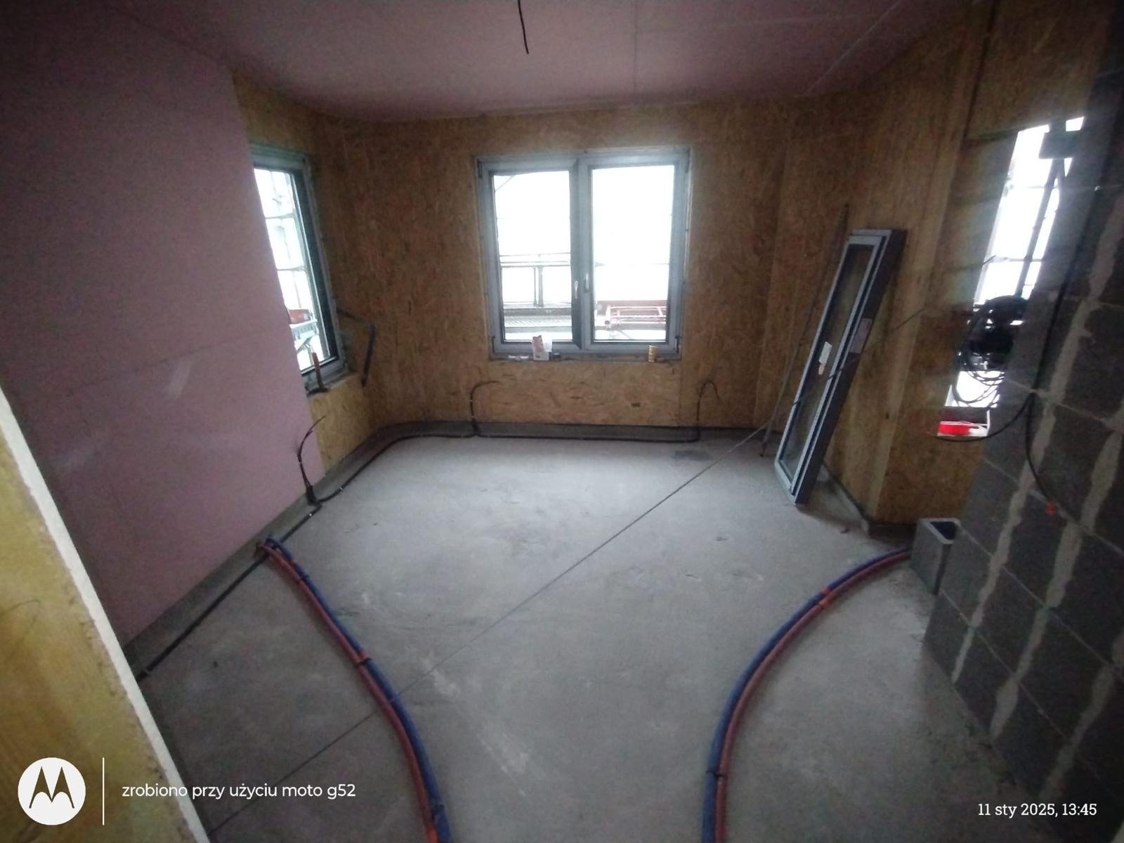 Wnętrze w trakcie budowy, ściany wyłożone płytami OSB, widoczne okna, instalacja grzewcza podłogowa częściowo ułożona, fragment ściany z bloczków betonowych.