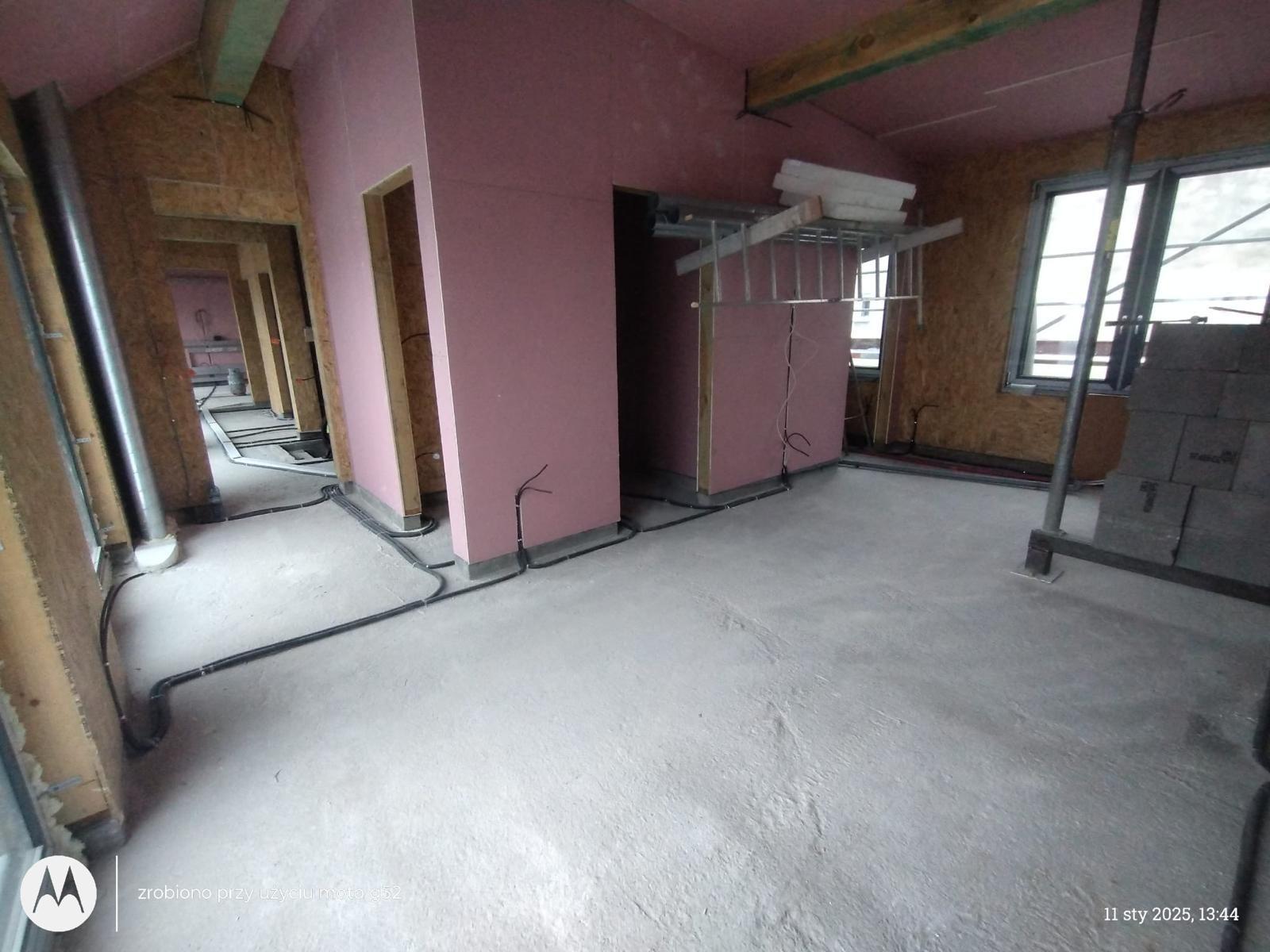 Wnętrze domu w trakcie budowy: widoczne ściany działowe z płyt gipsowo-kartonowych w kolorze różowym, surowa podłoga, rozprowadzone kable elektryczne w listwach przypodłogowych oraz drewniane...