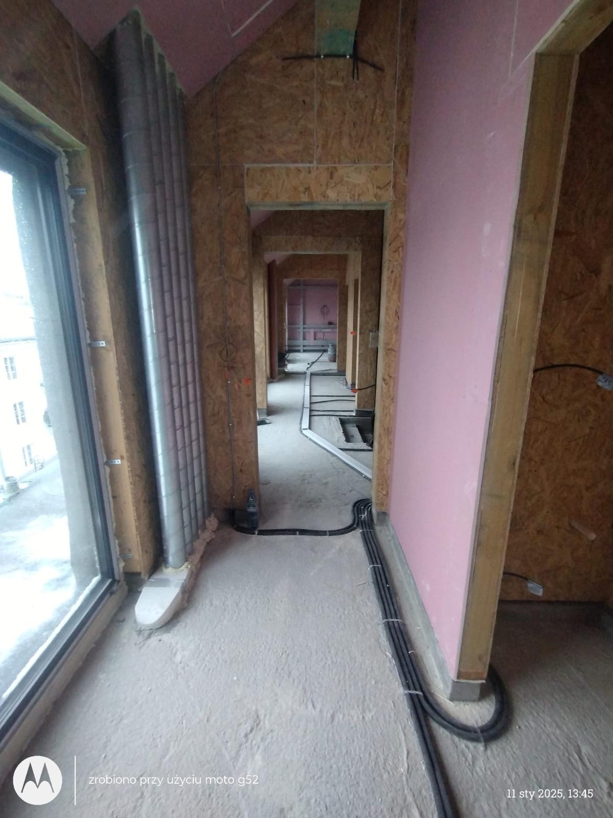 Wnętrze domu w stanie surowym: widok na korytarz z widocznymi instalacjami elektrycznymi, wentylacyjnymi, ścianami z płyt OSB i częściowo wykończonymi płytami gipsowo-kartonowymi.