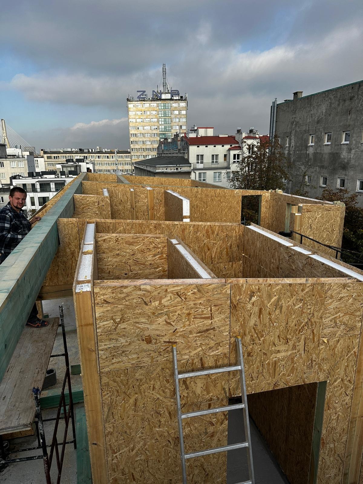 Wznoszenie konstrukcji ścian szkieletowych z płyt OSB na dachu budynku w miejskiej scenerii, widoczna drabina i pracownik w kraciastej koszuli.