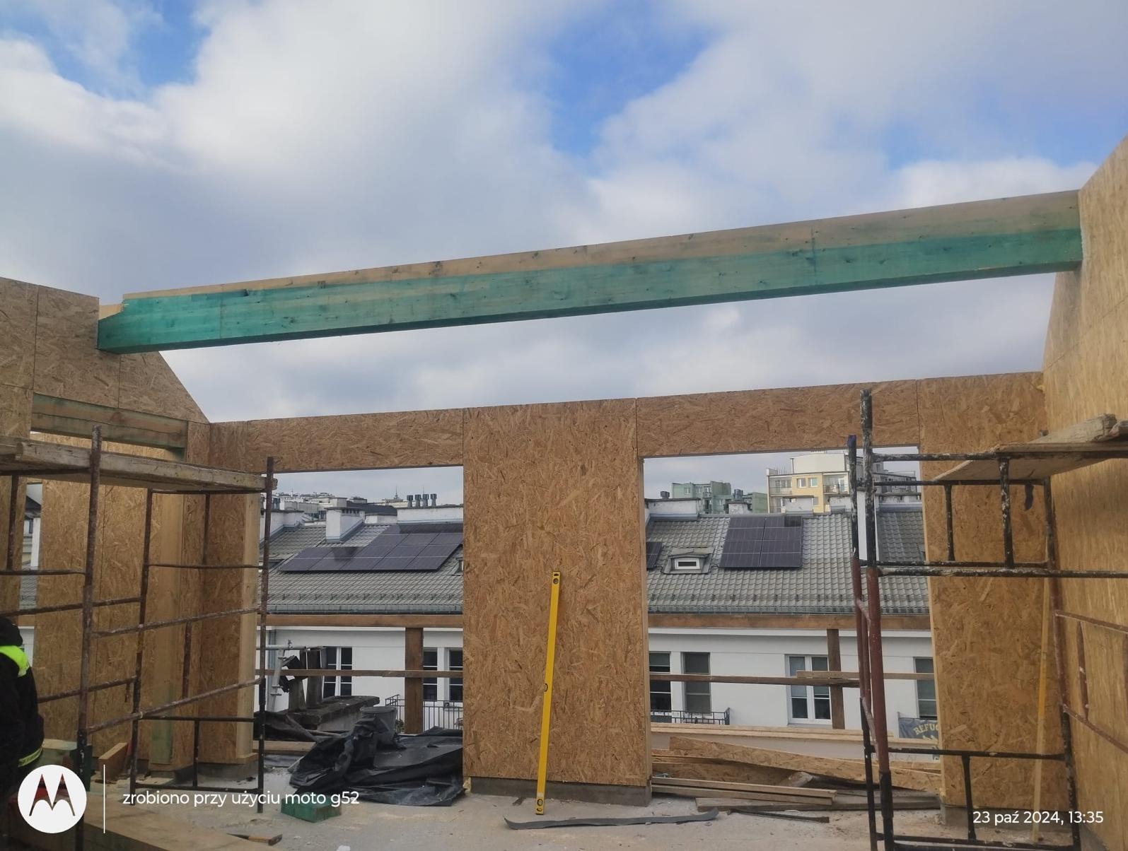 Wznoszenie ścian szkieletowych z płyt OSB w Wólce Radzymińskiej, widoczna poziomica i rusztowanie, w tle budynki z panelami słonecznymi.