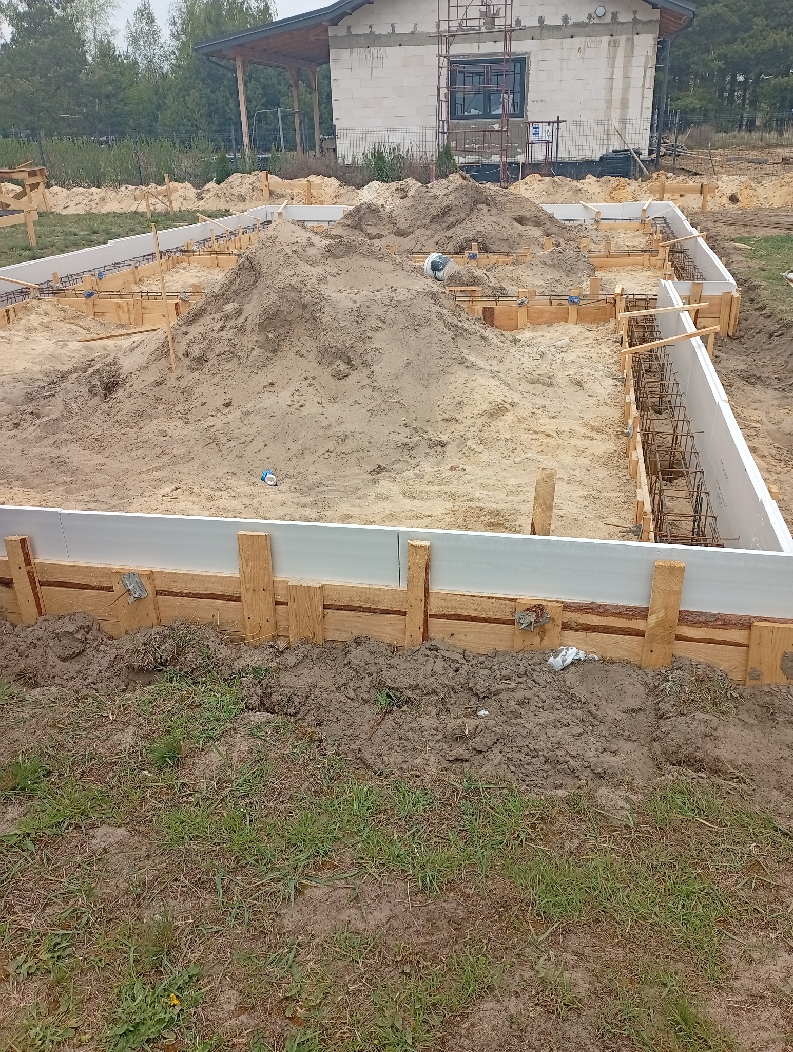 Fundamenty domu modułowego w trakcie budowy: szalunek z drewnianych desek, zbrojenie, nasypany piasek. W tle budynek w stanie surowym otwartym.