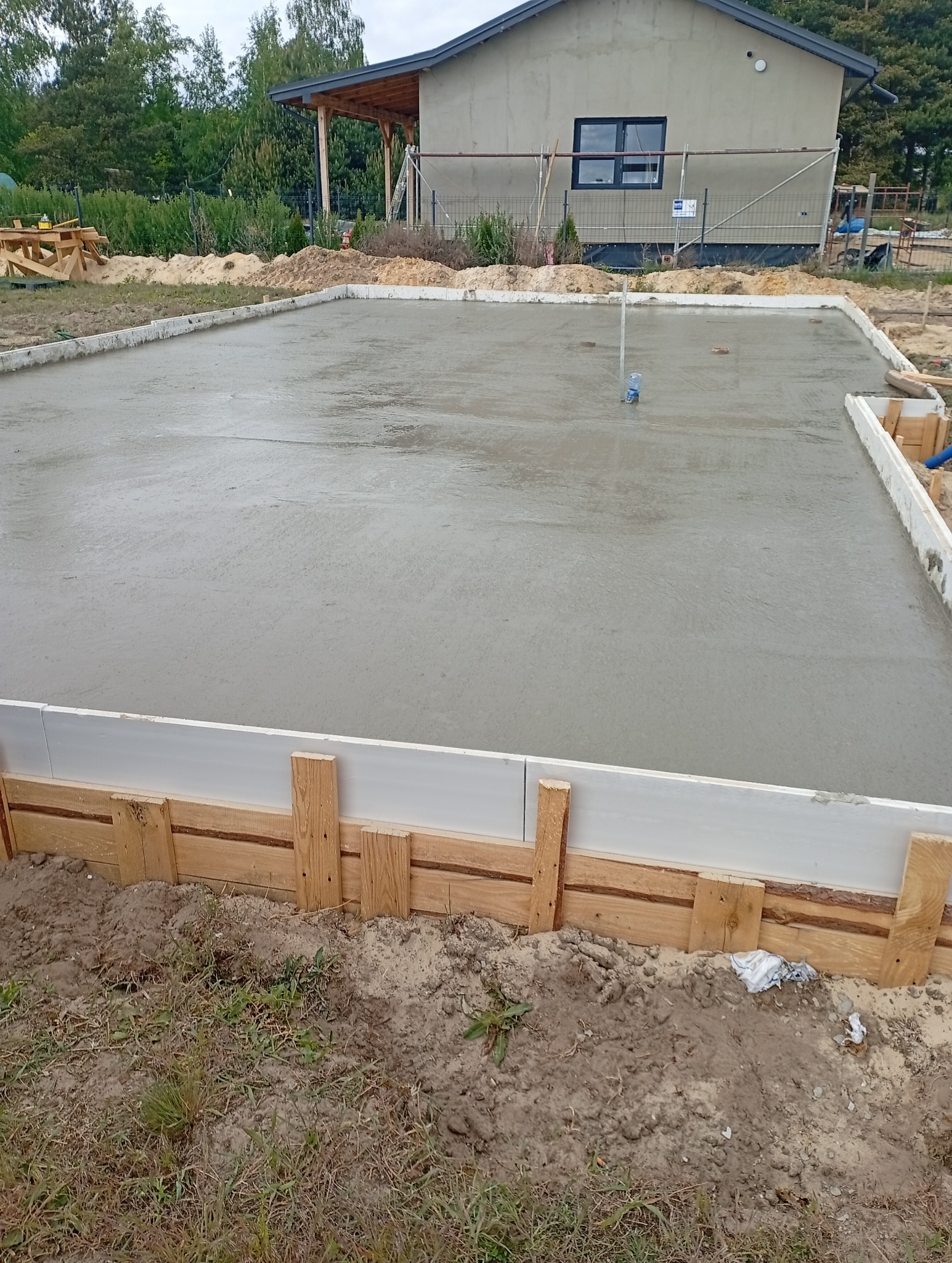 Wylany fundament pod dom modułowy z widocznym szalunkiem i elementami konstrukcyjnymi w tle, w otoczeniu zieleni i drzew. Etap budowy.