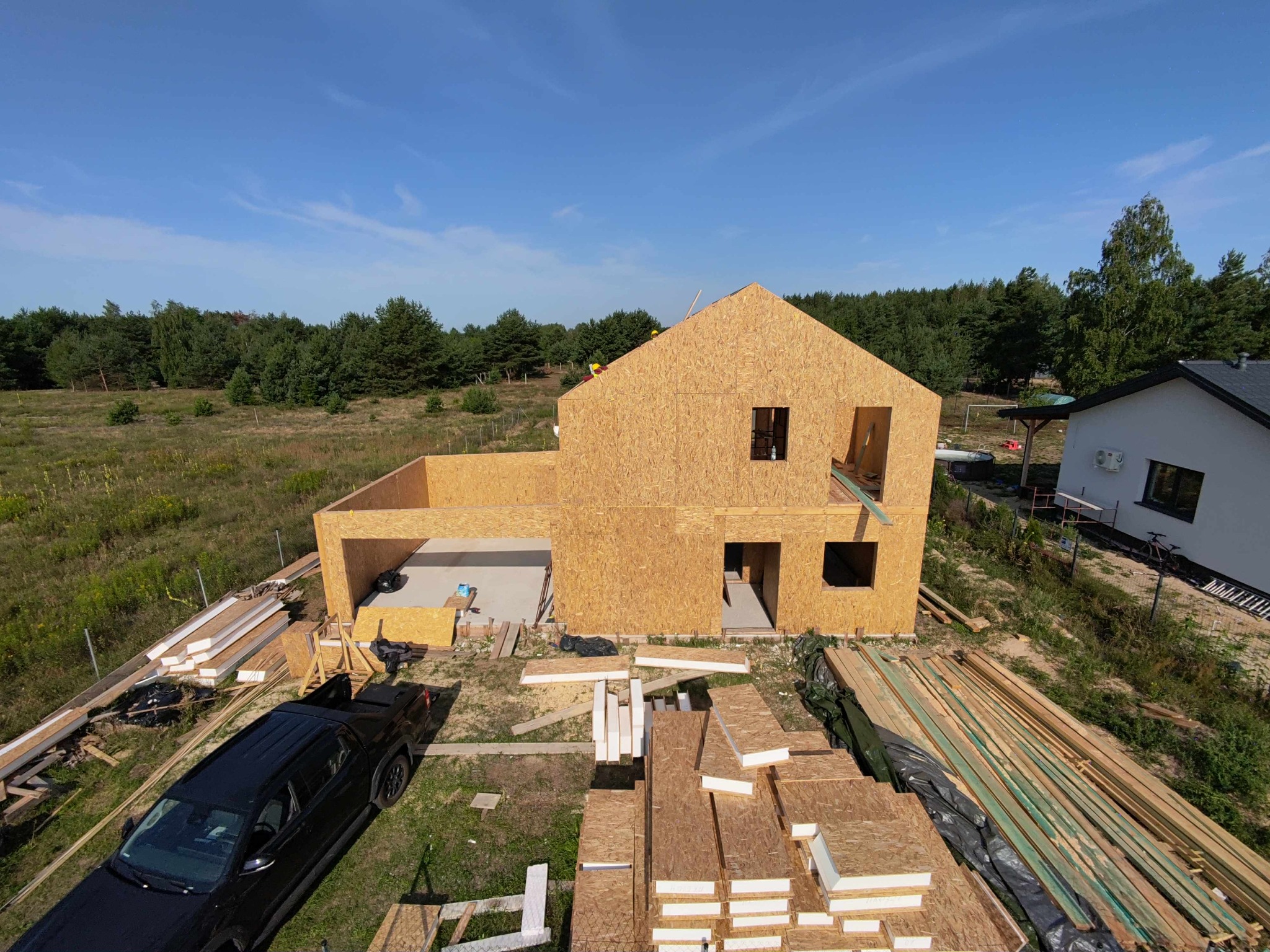Wznoszenie szkieletu domu z płyty OSB na tle lasu i błękitnego nieba. Widoczne elementy konstrukcyjne, narzędzia i materiały budowlane, czarny samochód.