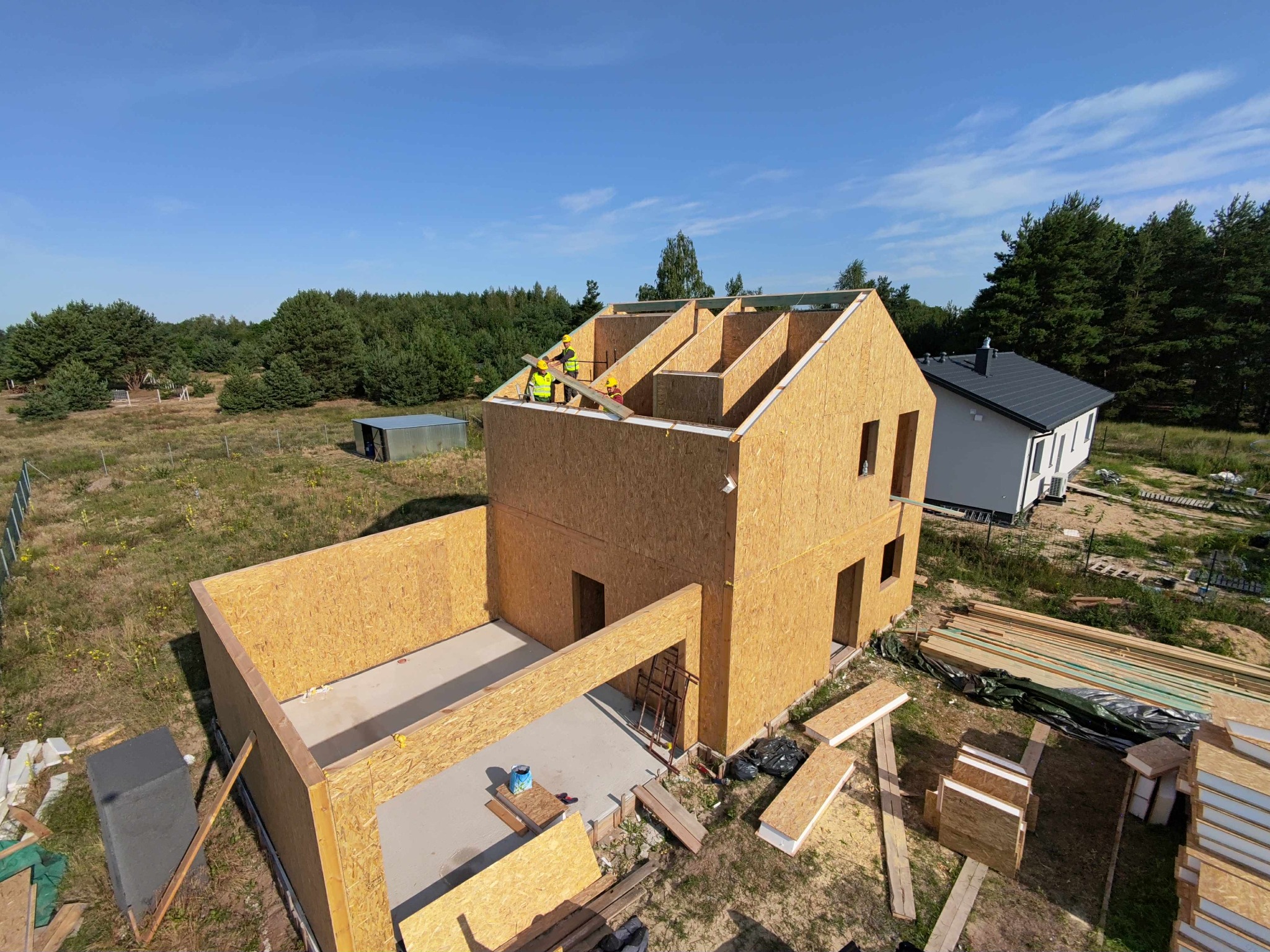 Budowa domu modułowego w trakcie: montaż ścian i dachu przez robotników w kaskach. Widok z góry na plac budowy z elementami konstrukcyjnymi i zielonym tłem.