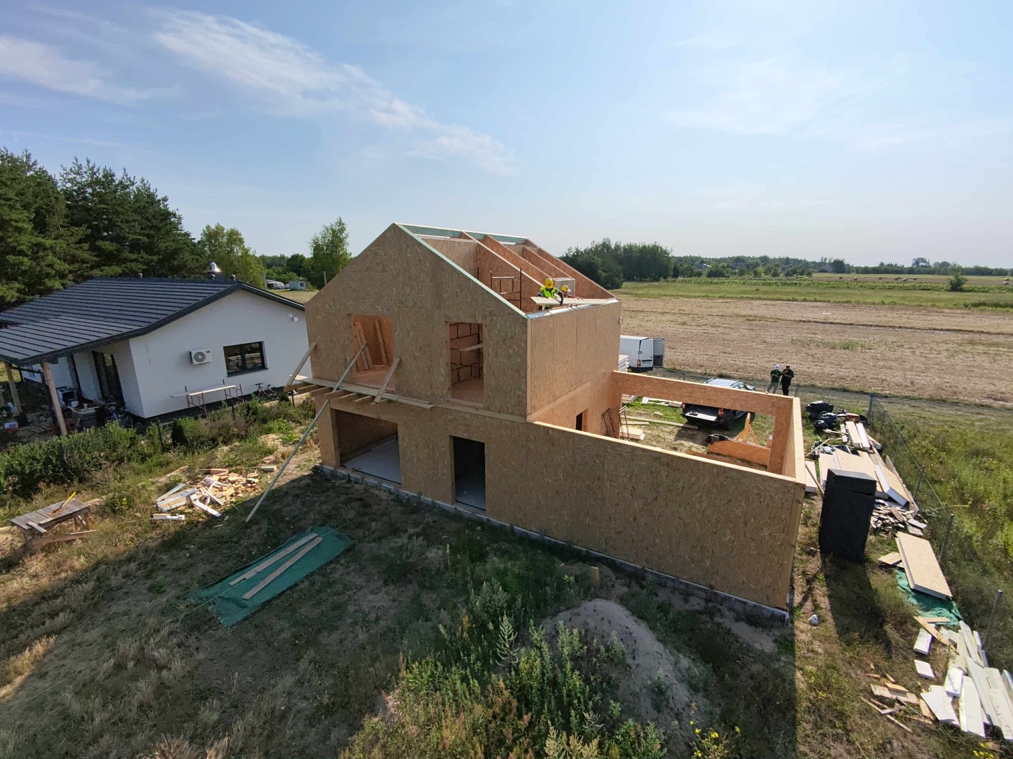 Wznoszenie szkieletu domu z płyt OSB na fundamencie, widok z góry. Dalszy plan: dom z ciemnym dachem i pole. Dwóch pracowników w oddali.