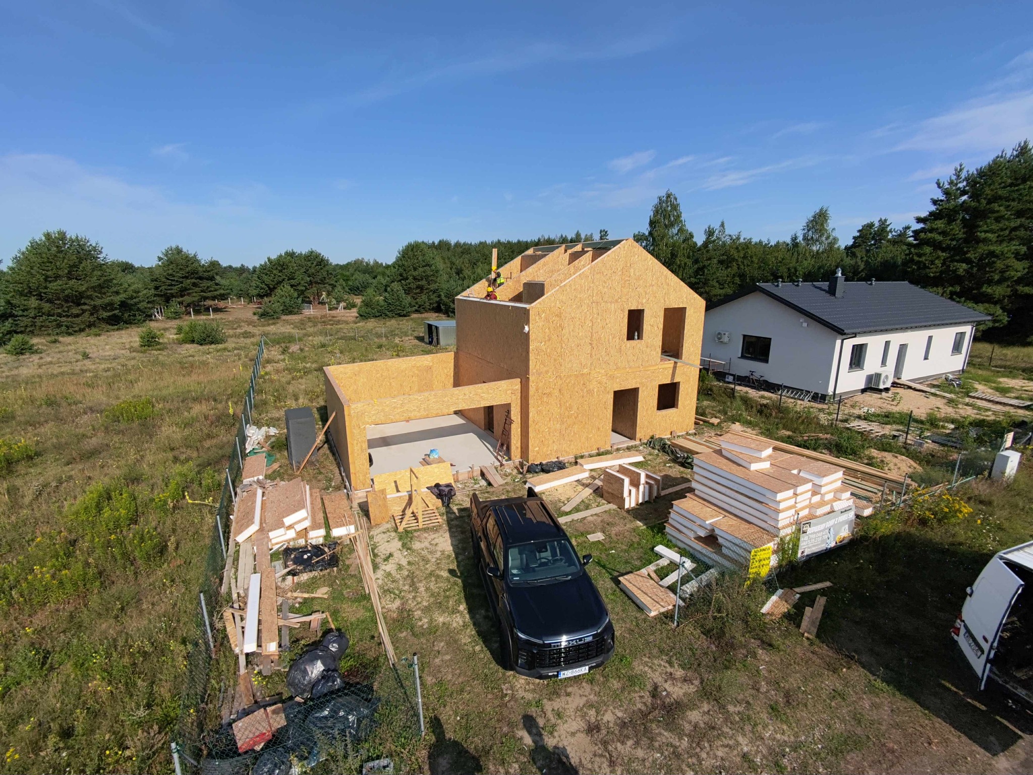 Budowa domu w Wólce Radzymińskiej: etap montażu szkieletu z OSB. Widoczny samochód, materiały budowlane i fragment gotowego domu obok.