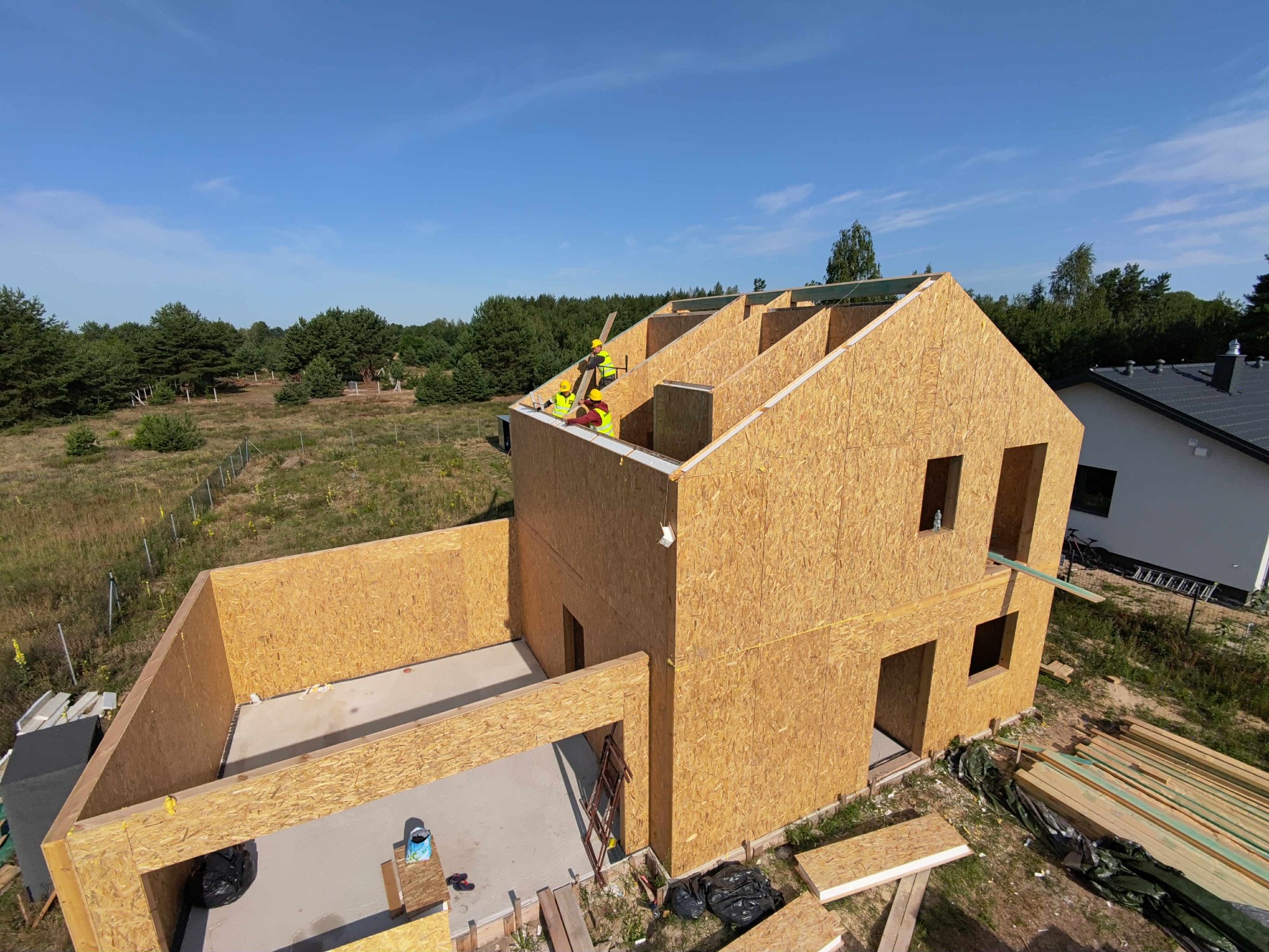 Budowa domu z drewnianych paneli OSB. Widoczne fragmenty konstrukcji, pracownicy w kaskach. W tle zieleń i błękitne niebo. Ujęcie z góry, widać postęp prac.