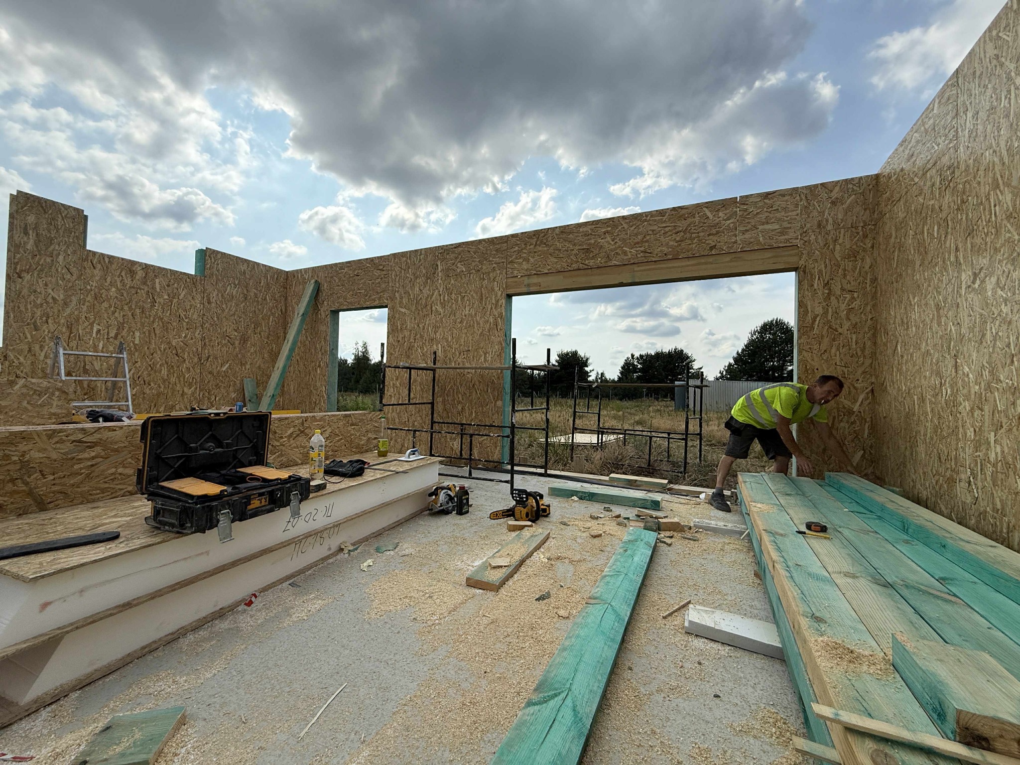 Montaż domu modułowego: pracownik w kamizelce układa zielone deski wewnątrz konstrukcji z płyt OSB, widoczne narzędzia i skrzynka narzędziowa.