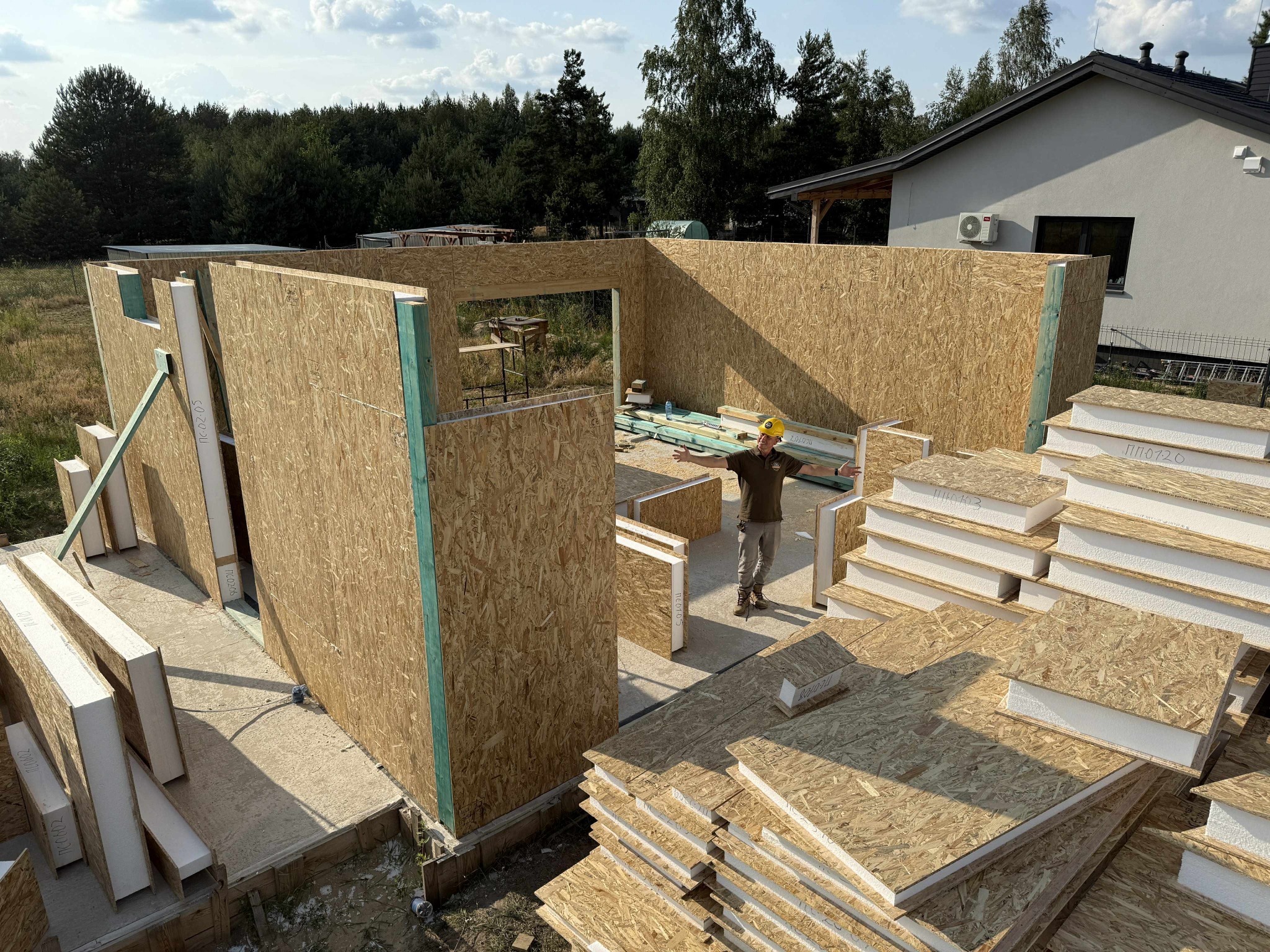 Wznoszenie ścian domu z paneli drewnianych. Na pierwszym planie stosy paneli izolacyjnych. W tle pracownik w kasku z rozłożonymi rękami.