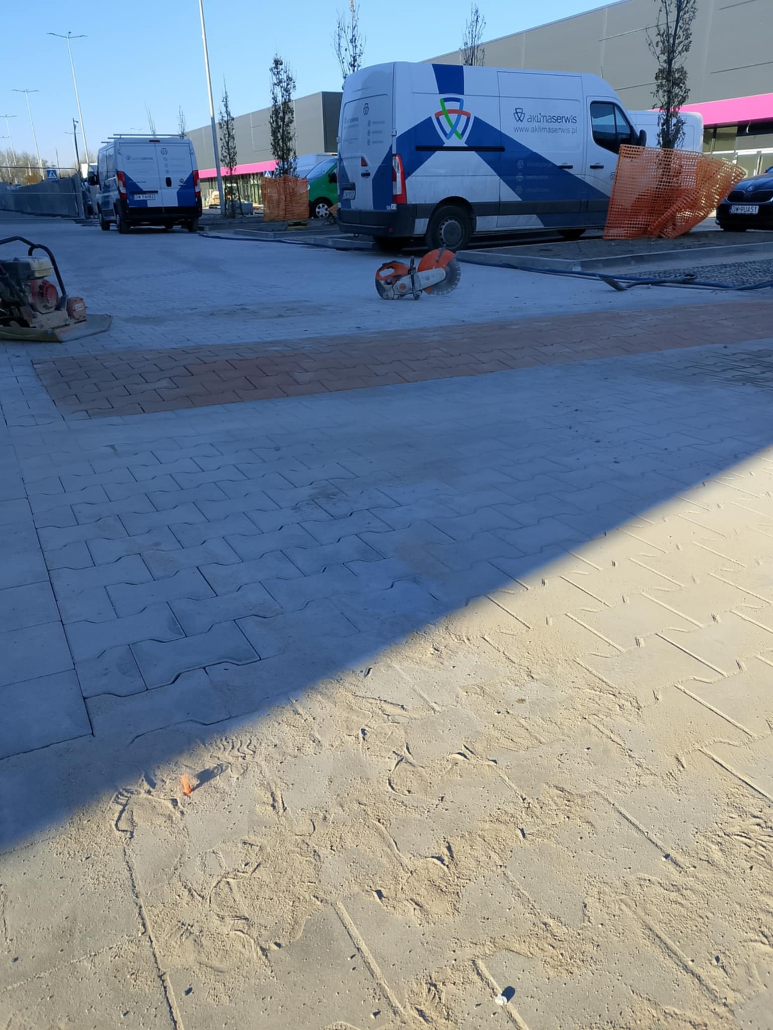 Układanie kostki brukowej na parkingu, widoczny fragment nowej nawierzchni, piasek, maszyna do ubijania, przecinarka do kostki i firmowe busy w tle.