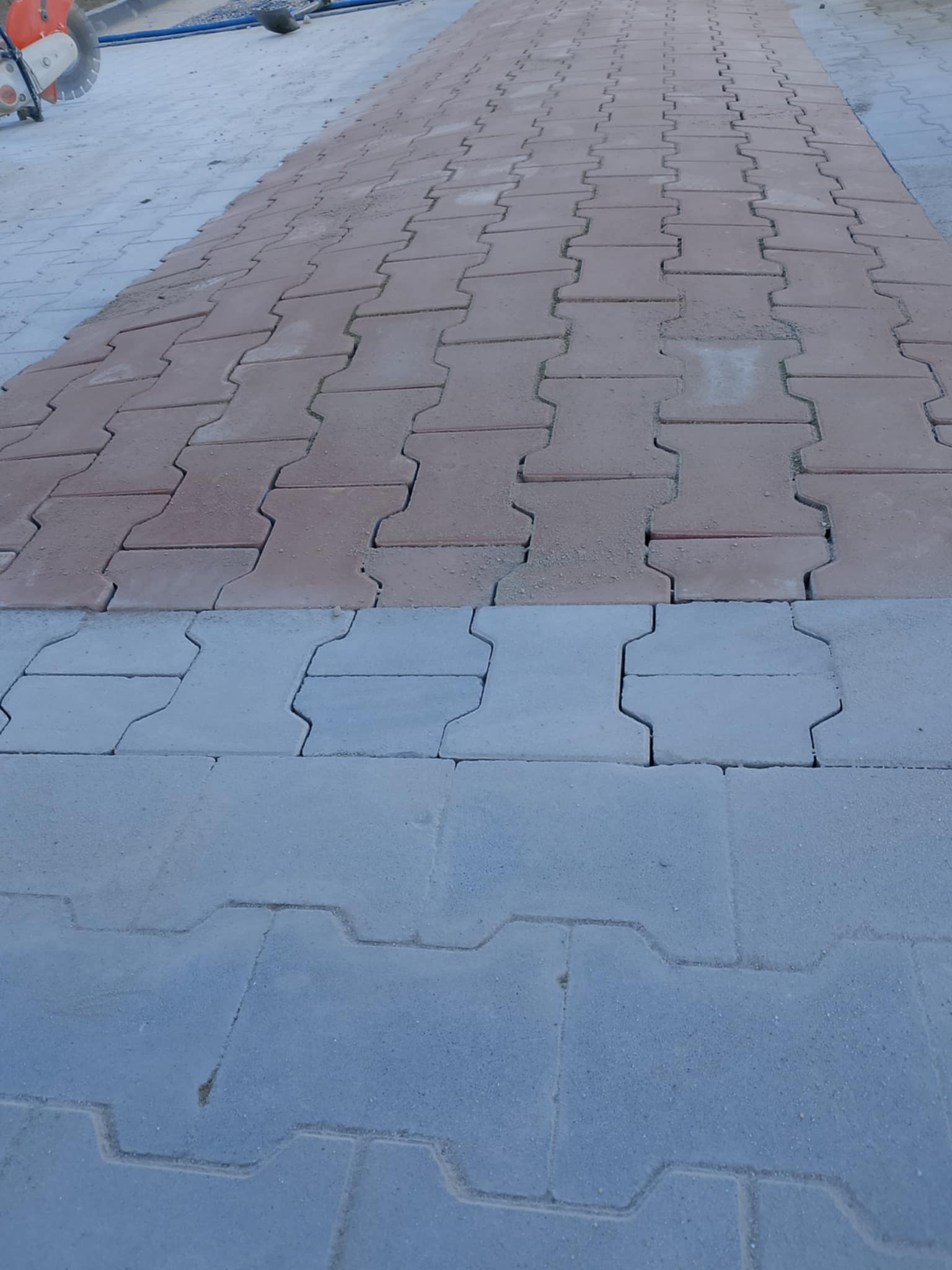 Ułożona, dwukolorowa kostka brukowa typu 'I': szara i czerwona, z widoczną przecinarką do kostki brukowej w lewym górnym rogu.