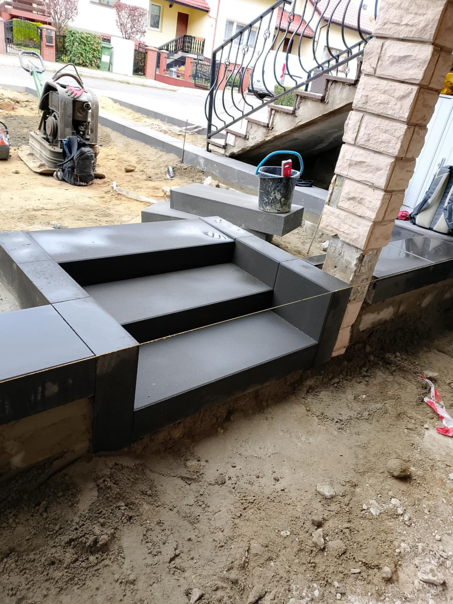 Montaż ciemnoszarych płytek na schodach zewnętrznych domu, widoczna ubijarka w tle i wiadro z zaprawą na górnym stopniu.