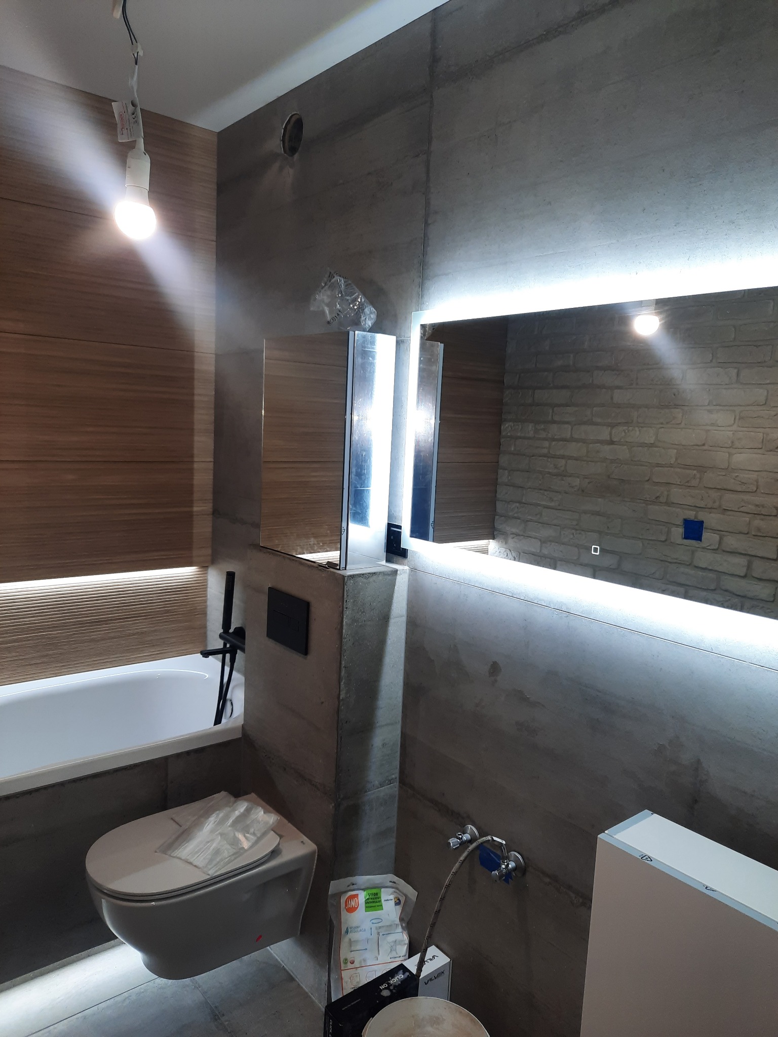 Nowoczesna łazienka w trakcie remontu: widoczna wanna, podwieszana toaleta z folią ochronną, ściany wykończone płytkami imitującymi beton i drewno, lustro z podświetleniem LED, wisząca żarówka...