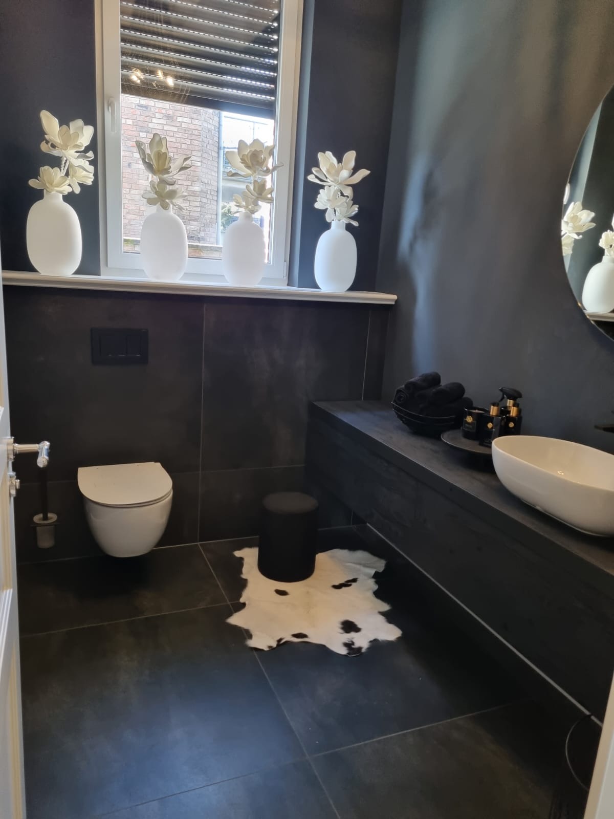 Nowoczesna łazienka w odcieniach szarości z czarnymi akcentami, białą ceramiką, czterema wazonami z białymi kwiatami na parapecie okna i dywanikiem ze wzorem krowich łat na ciemnej podłodze.