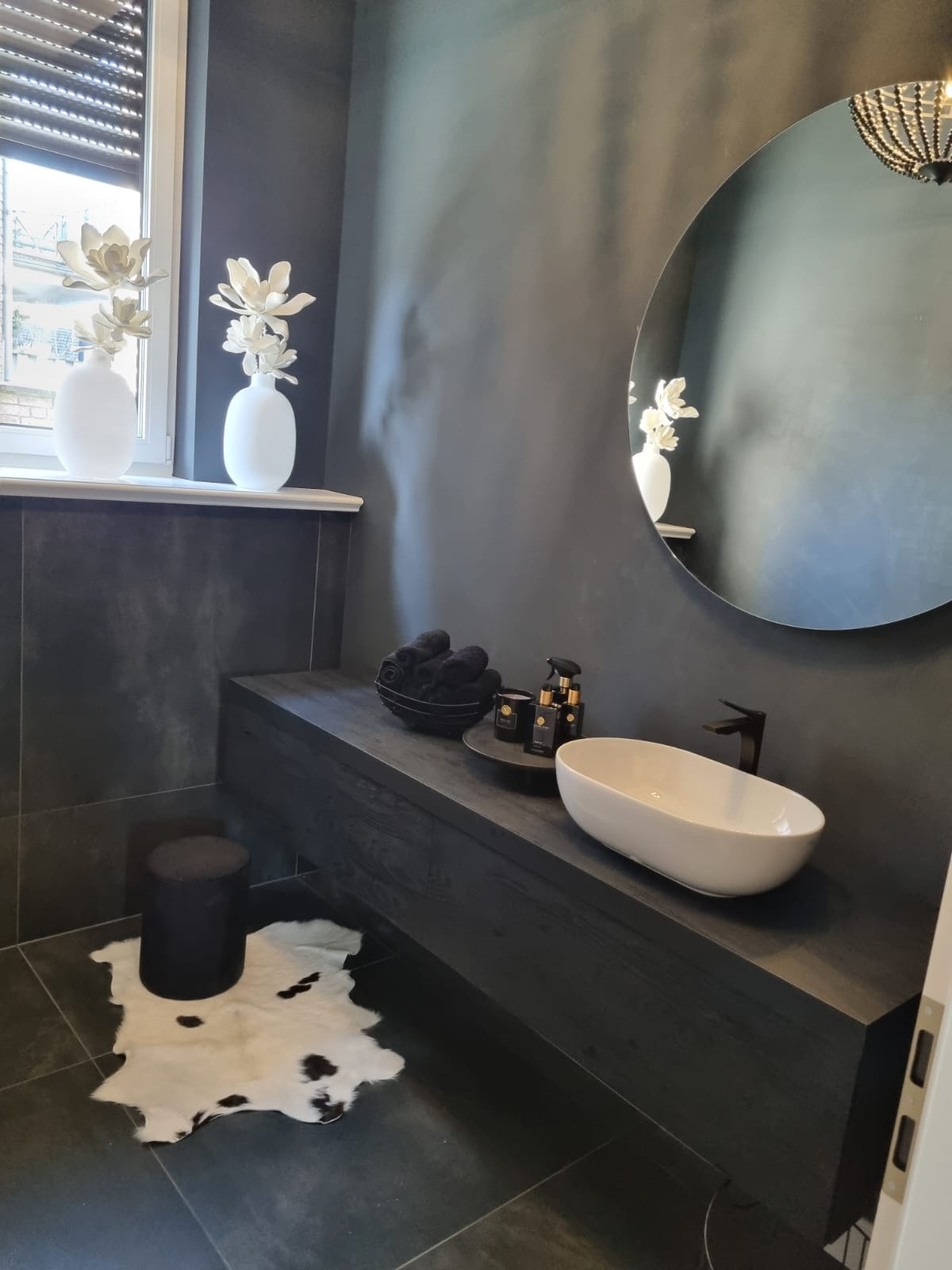 Nowoczesna łazienka w odcieniach szarości z owalną umywalką nablatową, okrągłym lustrem i dekoracyjnymi wazonami z kwiatami na parapecie okna, uzupełniona czarnymi ręcznikami i dywanikiem w cętki.