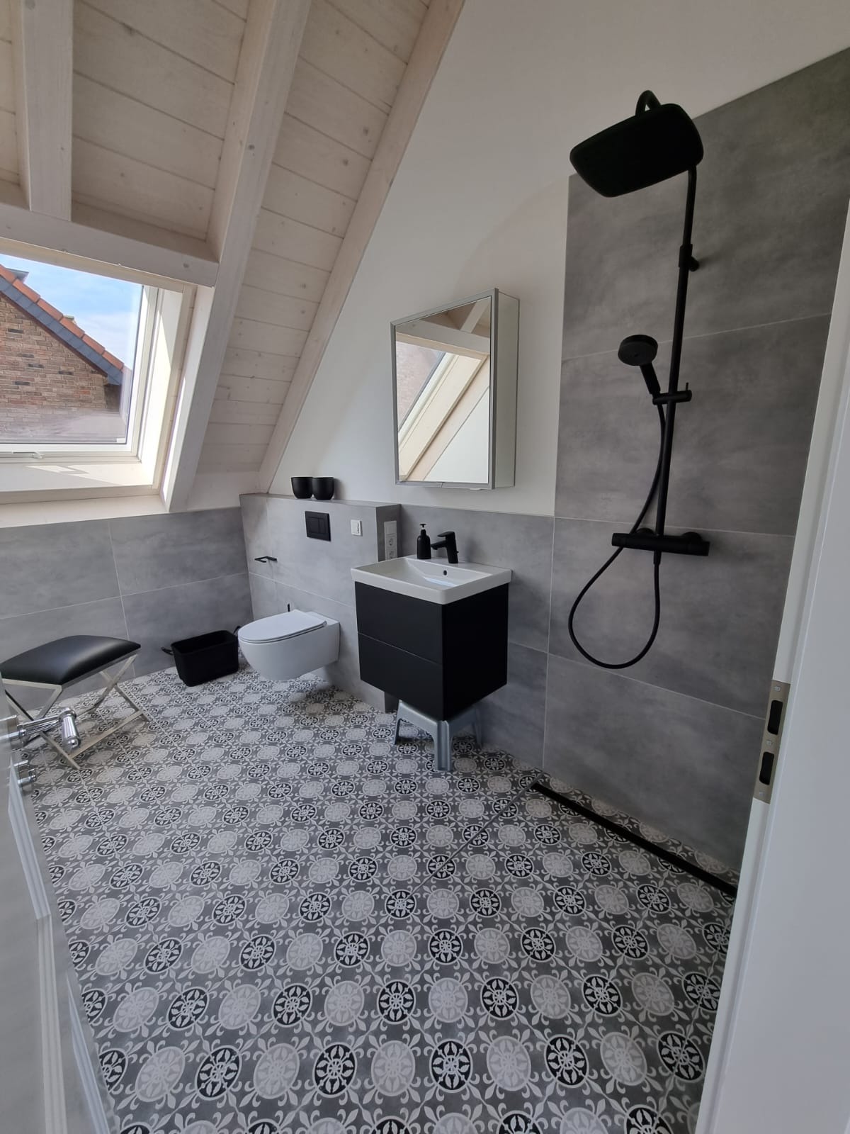 Nowoczesna łazienka na poddaszu z szarymi płytkami, czarną armaturą i wzorzystą podłogą, widok na okno dachowe.
