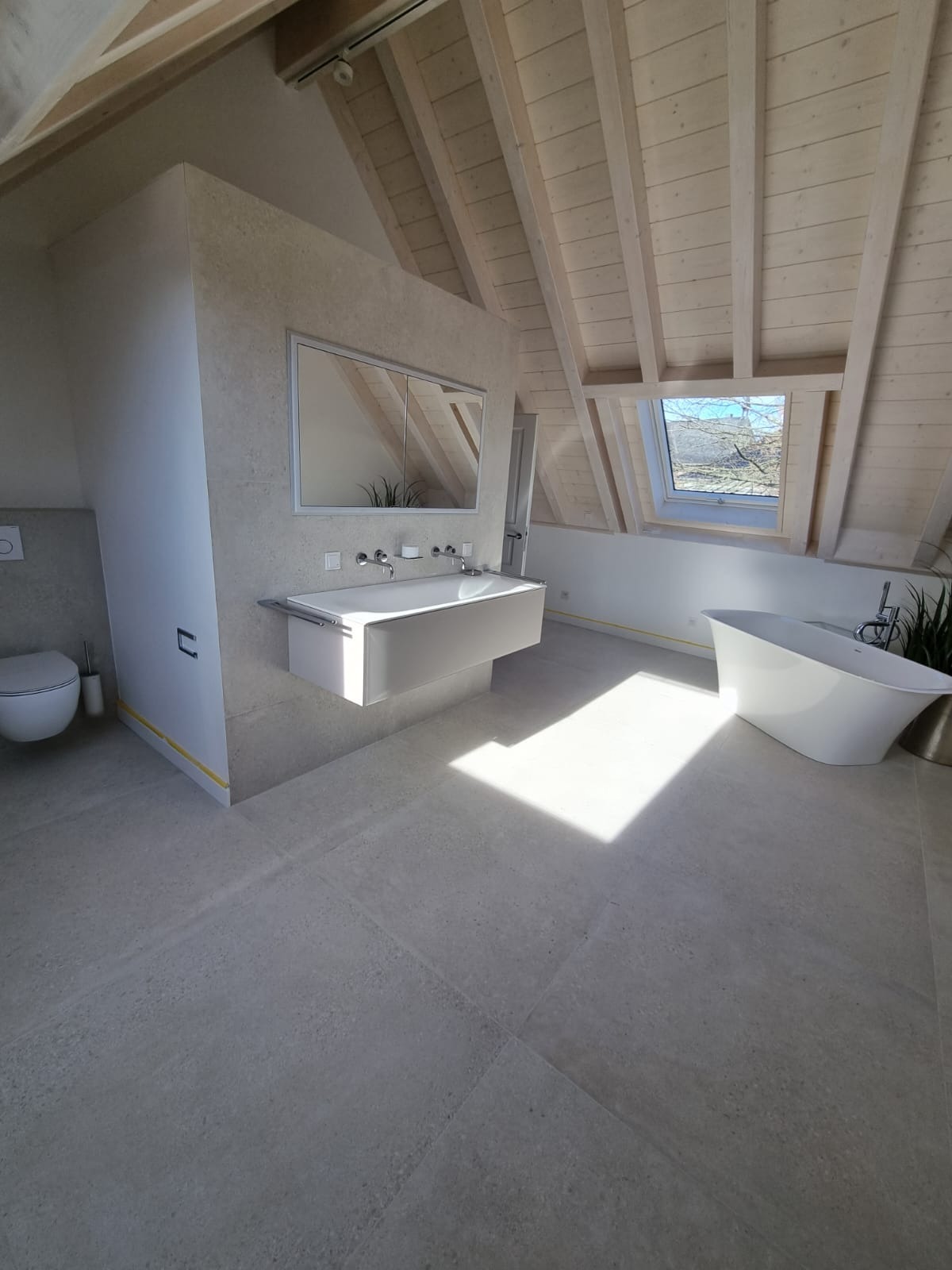 Jasna łazienka na poddaszu z drewnianym stropem, wolnostojącą wanną i podwieszaną umywalką z szafką lustrzaną, wpadające światło przez okno dachowe.