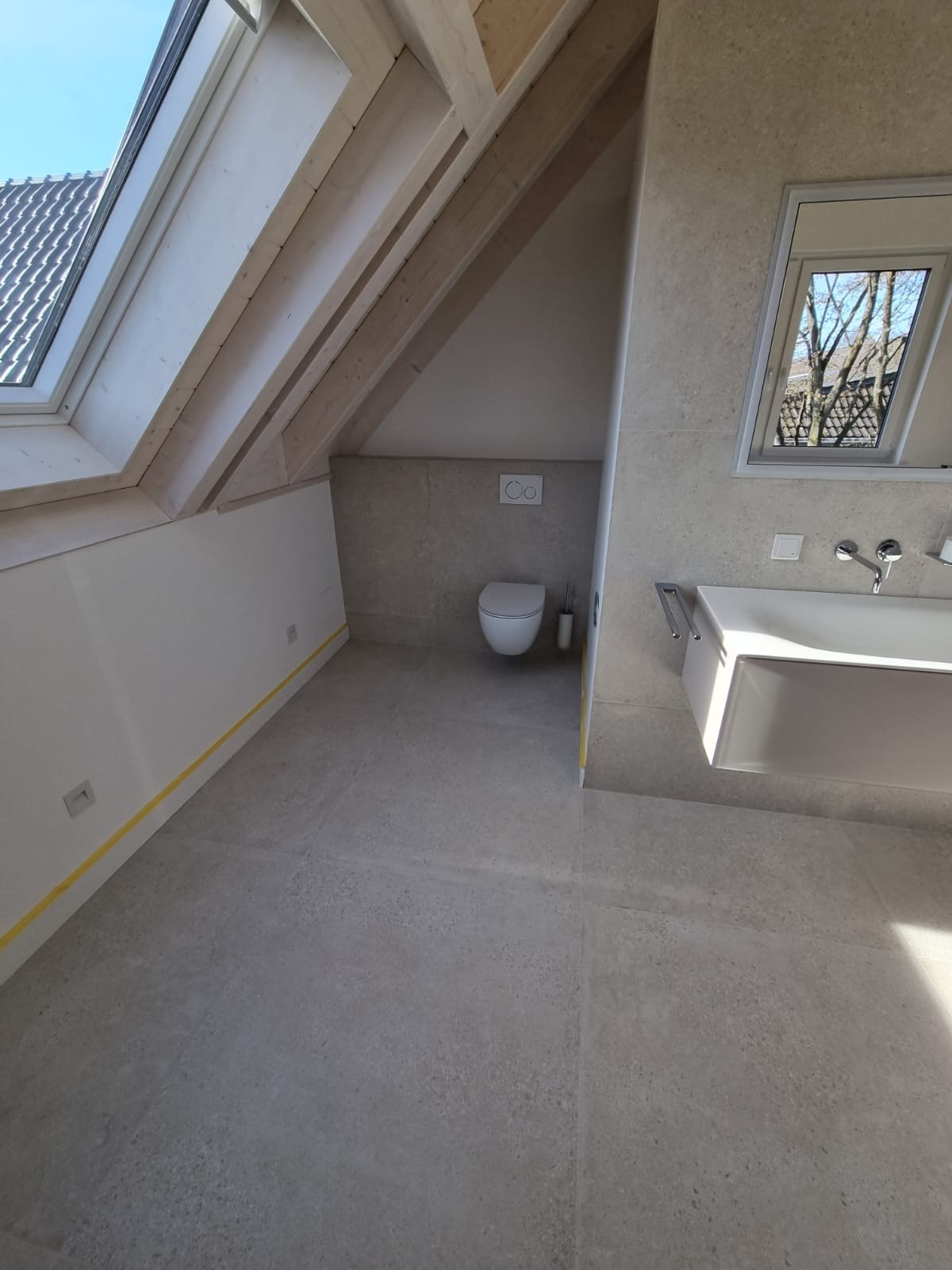 Jasnoszara łazienka na poddaszu z oknem dachowym, widoczna toaleta, umywalka i fragment drewnianej konstrukcji dachu, zabezpieczona taśmą malarską przy podłodze.