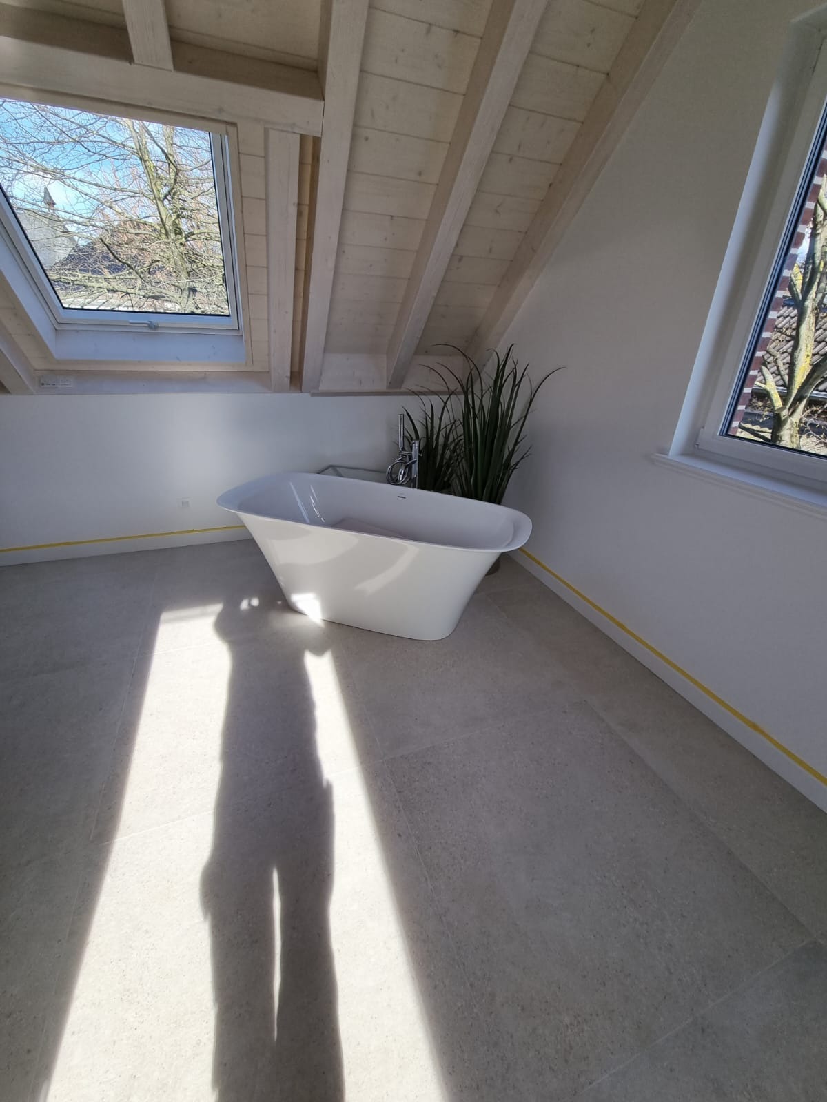 Wolnostojąca wanna w jasnej łazience na poddaszu z oknami dachowymi, widoczna cień osoby robiącej zdjęcie, żółta taśma malarska przy podłodze.