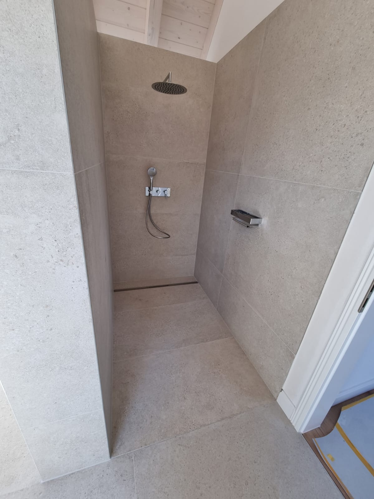 Kabina prysznicowa wykończona beżowymi płytkami imitującymi kamień, z odpływem liniowym, deszczownicą i słuchawką prysznicową na ścianie, widok z góry.