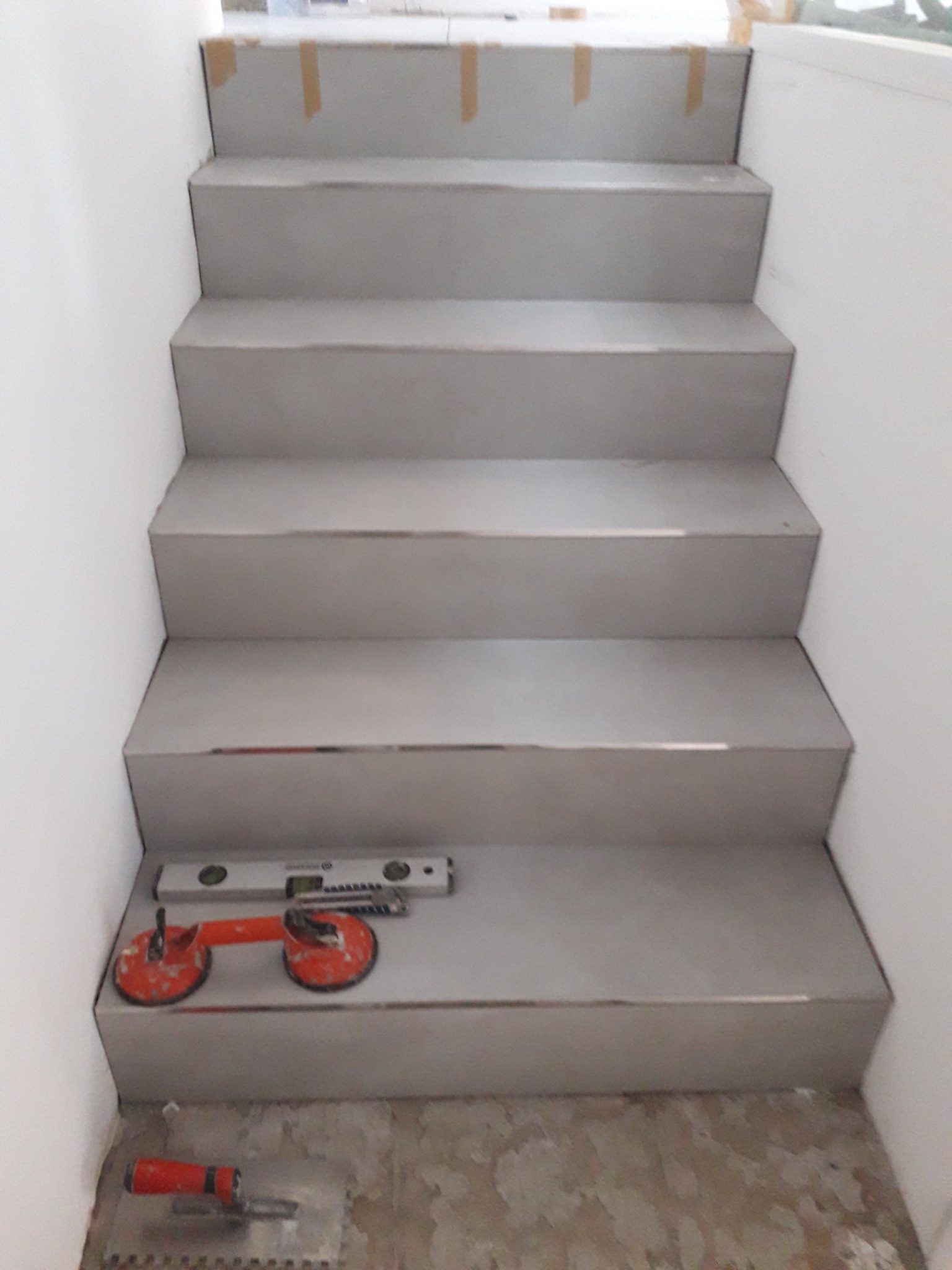 Wykończone schody z szarych płytek, poziomica i przyssawki do płytek na jednym ze stopni. Widoczne narzędzia glazurnicze na podłodze.